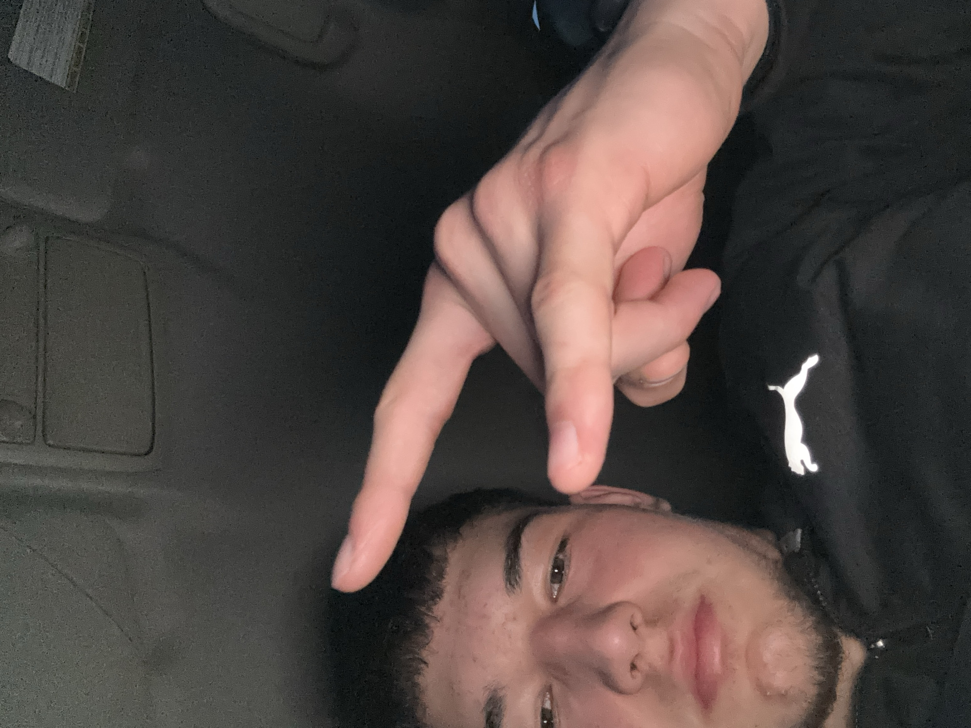 A person lies on their back in an upholstered car interior. The person is wearing a black Puma jacket. One hand is raised, making a "V" sign over the person's face. A portion of the car's headliner is visible. The location cannot be determined from the image.