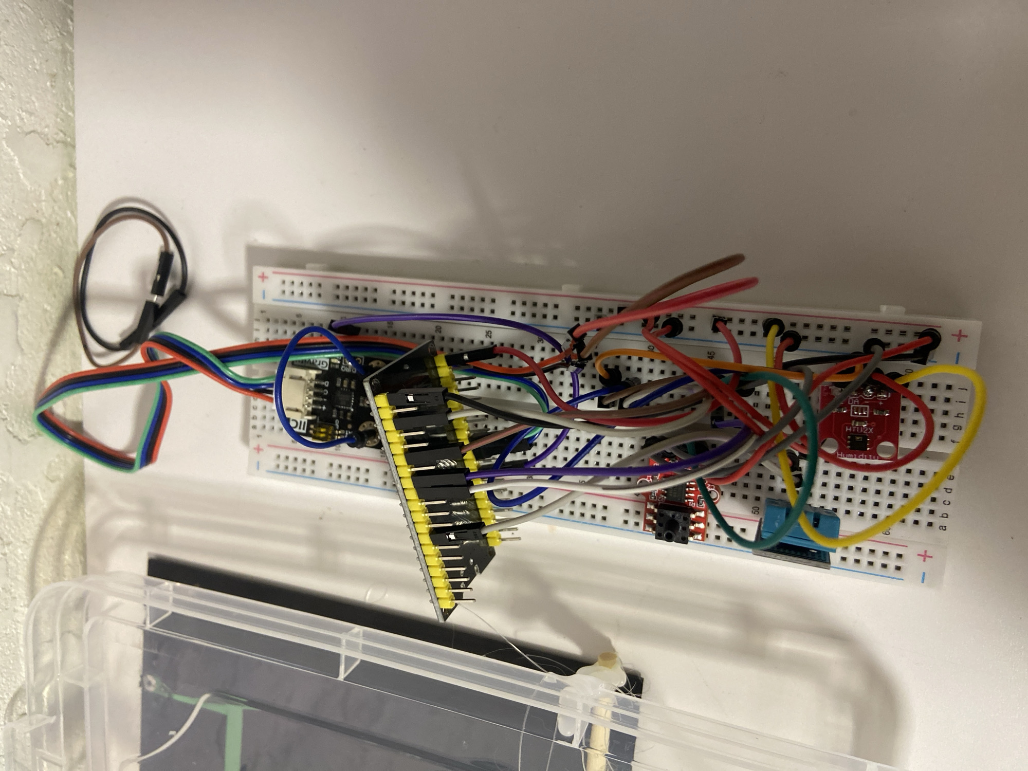In an indoor, likely home or workshop setting, a white table surface serves as the backdrop for a complex electronic circuit experiment. The scene is dominated by a white breadboard, a common prototyping tool for electronics, adorned with a multitude of colorful jumper wires. These wires, red, blue, green, purple, gray, yellow, and brown, connect various components.
Prominently featured on the breadboard are several integrated circuits and modules. A black circuit board with yellow pin headers is positioned towards the top, with wires emanating from it. Below this, a smaller red circuit board with a black button is visible. Further down, a blue component with a black header is integrated into the circuit. Additionally, a small red module with the text "Humiditi" and "HTU2X" is present.
To the left of the breadboard, a clear plastic container or lid is partially visible, suggesting a workspace or storage area for electronic components. A few strands of white string or wire are also present near this container. The overall impression is one of an active electronics project, with the various components and wiring indicating a functional, albeit potentially unfinished, circuit. The lighting suggests artificial indoor illumination, casting subtle shadows.