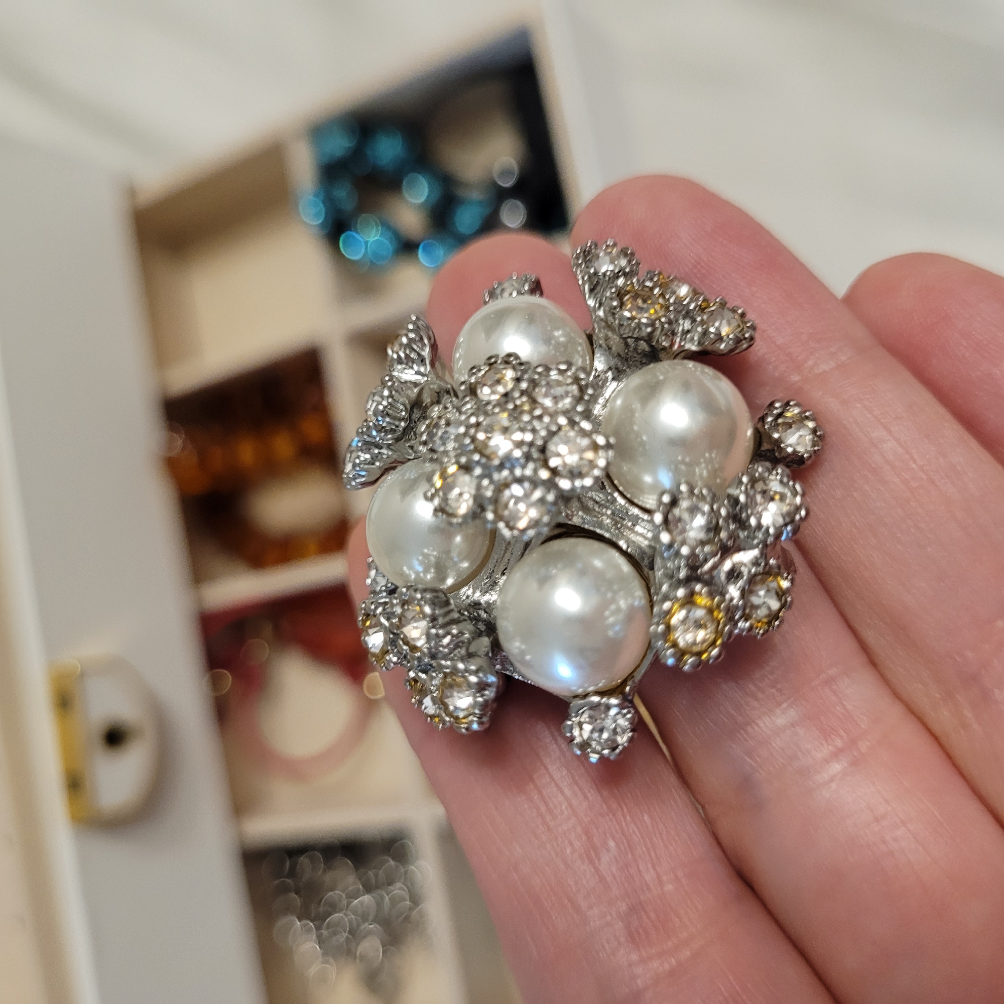 A close-up, high-angle shot captures a hand gently holding an ornate piece of jewelry against a softly blurred background. The jewelry, likely a ring or brooch, features a central cluster design comprising five prominent white pearls, surrounded and interspersed with numerous sparkling rhinestones of varying sizes. Most rhinestones appear clear or silver-toned, while a few have a subtle yellowish tint, all set in a reflective silver-colored metal. The light catches the pearls, highlighting their smooth, iridescent surfaces, and makes the rhinestones gleam.
The hand, with visible skin texture and faint lines, holds the jewelry, allowing its intricate details to be the focal point. In the out-of-focus background, a multi-compartment white jewelry box or organizer is visible. Within its sections, several other pieces of jewelry can be seen as soft, vibrant blurs of color and light, creating a bokeh effect. These include bright blue items in the upper section and warmer, possibly orange or amber-toned items in a lower section, indicating a collection of diverse accessories. The scene suggests an indoor setting, perhaps a dressing area or bedroom, where jewelry is being examined or prepared for use. There is no visible text.