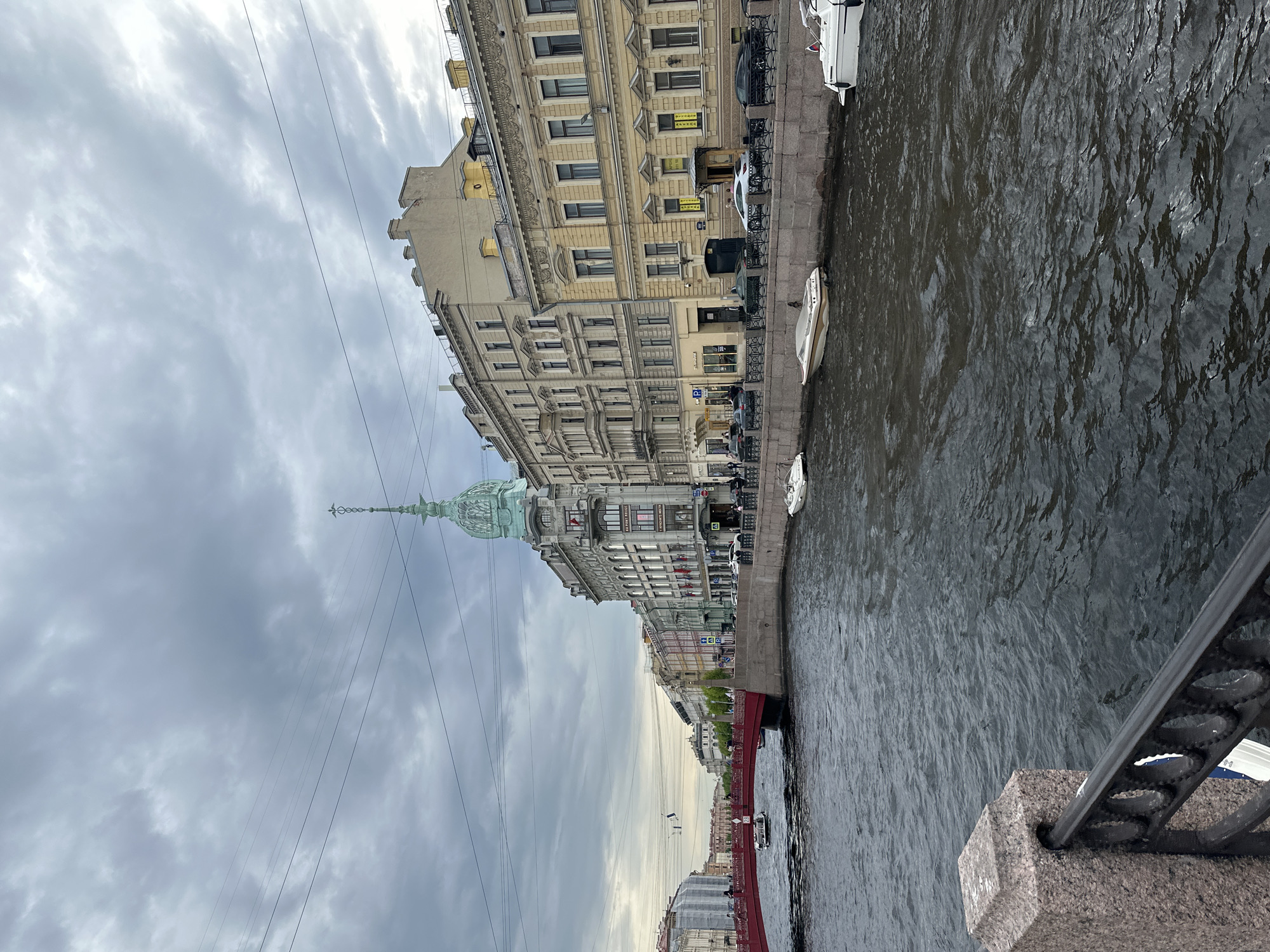 This image depicts a waterfront scene in Saint Petersburg, Russia. A wide river fills the right side of the image, its surface ruffled by wind or current. A stone embankment runs along the river's edge, with several small boats moored to it. Buildings, primarily of European architectural style, line the opposite side of the embankment, extending into the distance. The buildings vary in height and color, some with ornate facades. Overhead electrical lines crisscross the sky. The sky is overcast with grey clouds. In the middle ground, a bridge with red accents spans the river. The photo is taken from a slightly elevated viewpoint, looking out from a bridge or similar structure, with a portion of the bridge's railing visible in the lower right corner of the frame.