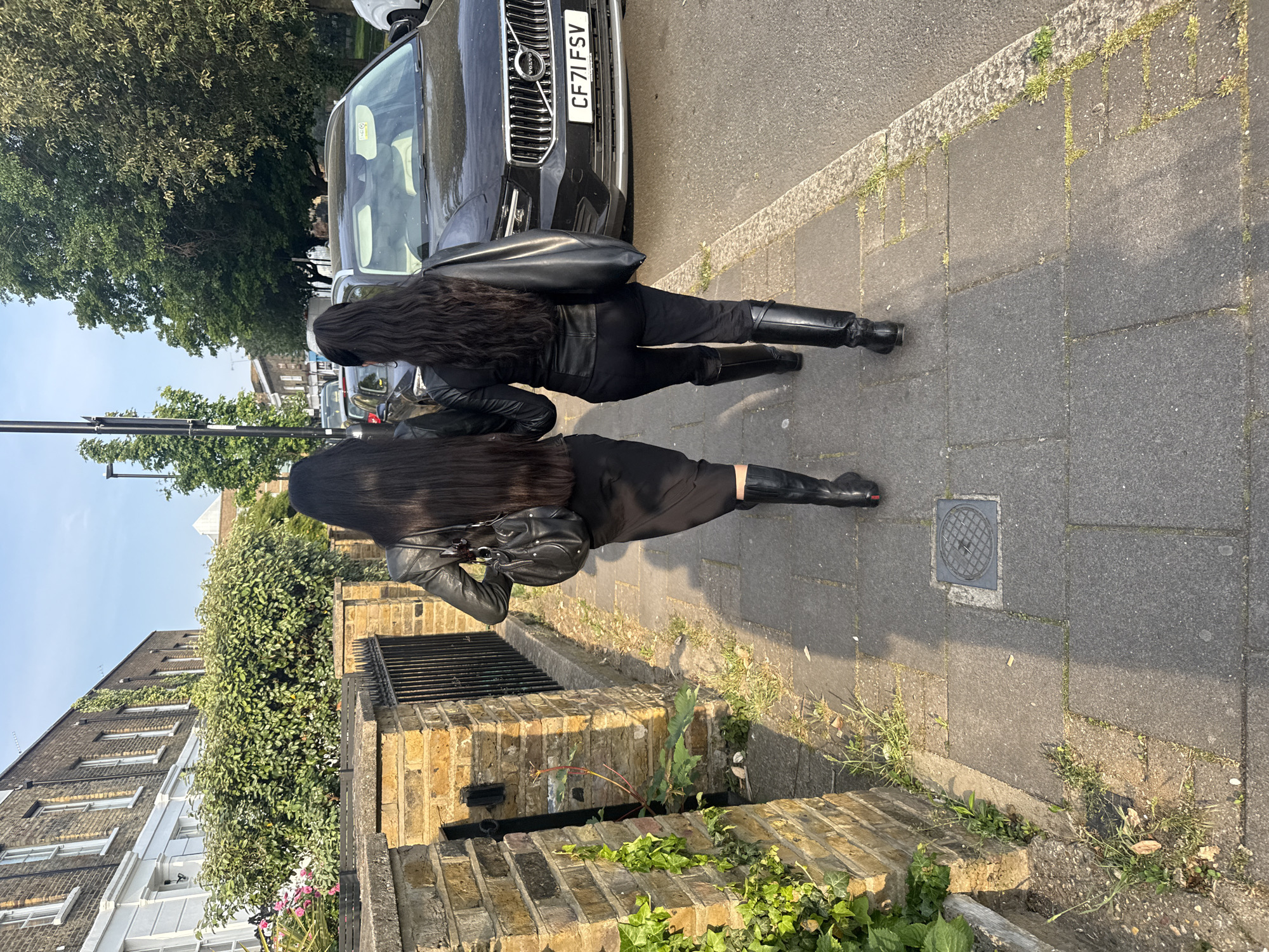 Two women with long dark hair, wearing black leather jackets, black pants or skirts, and black riding boots, walk side-by-side down a sidewalk. Each carries a black shoulder bag. A black Volvo SUV with license plate "CF71 FSV" is parked along the sidewalk. The scene is set in a residential area of London, United Kingdom, with brick buildings and greenery. The sidewalk is paved with gray stone.