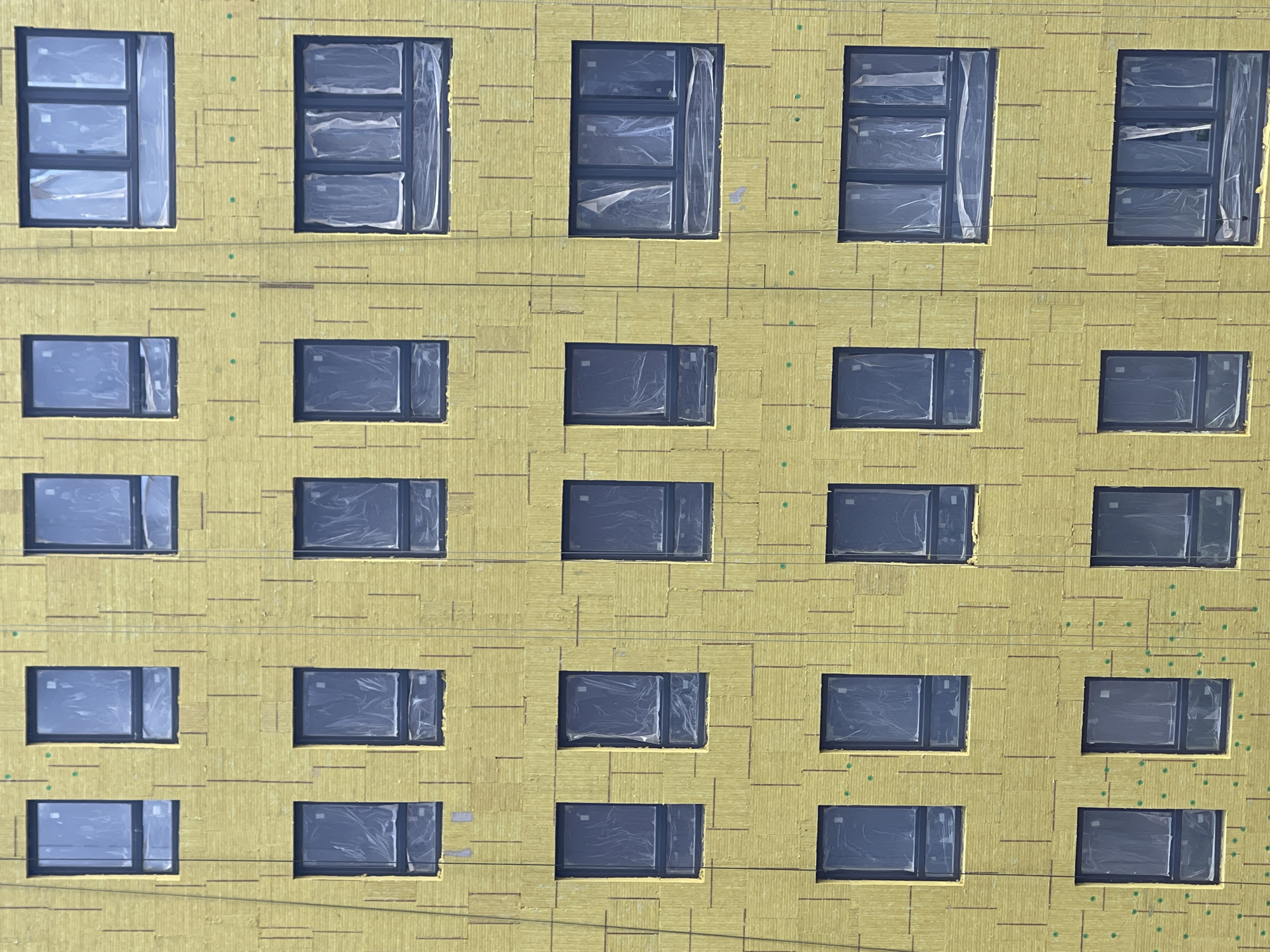 An exterior wall of a building under construction is visible, covered in yellow insulation panels. There are multiple windows of various sizes and configurations evenly spaced across the wall. The windows appear to be covered with a translucent plastic film, obscuring the view into the building. Thin vertical lines, possibly supporting wires or construction guides, are also present on the insulation. The overall impression is a modern building facade in a state of completion. The image does not contain people or any specific identifying landmarks that would indicate its location within a city.