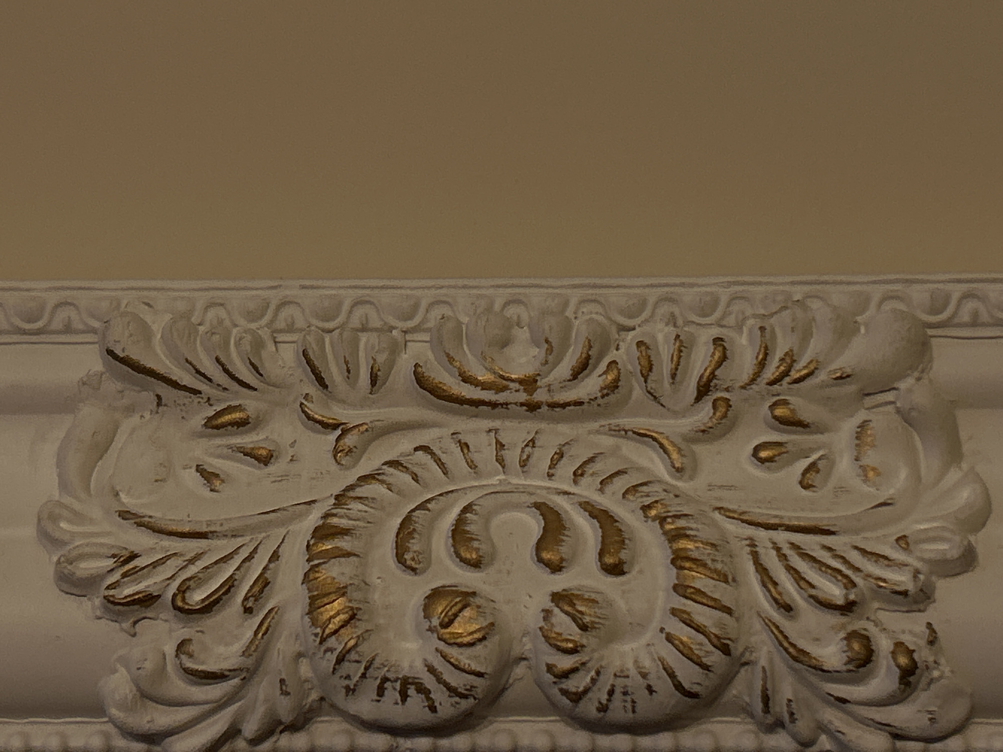 The image displays a close-up of an ornate architectural or furniture molding, possibly from an interior setting in Plovdiv, Bulgaria. The primary subject is a decorative relief carving, predominantly off-white or cream in color, with intricate details highlighted by metallic gold paint.
The molding features a symmetrical design comprising elaborate scrolls, volutes, and acanthus leaf-like motifs, characteristic of classical, Baroque, or Rococo revival styles. A larger, central, circular-like scroll pattern anchors the lower half, flanked by smaller, flowing scrollwork and foliage. Along the top edge of the main decorative element, a smaller, repetitive wavy or scalloped pattern is visible. The three-dimensional depth of the carving is enhanced by soft shadows. The painted surface shows signs of age or intentional distressing, with the white paint appearing slightly worn and the gold accents providing an antique or gilded effect. The background consists of a plain, solid-colored wall in a warm beige or light brown hue. No people, visible activities, events, or text are present in the frame. The lighting suggests an indoor environment.