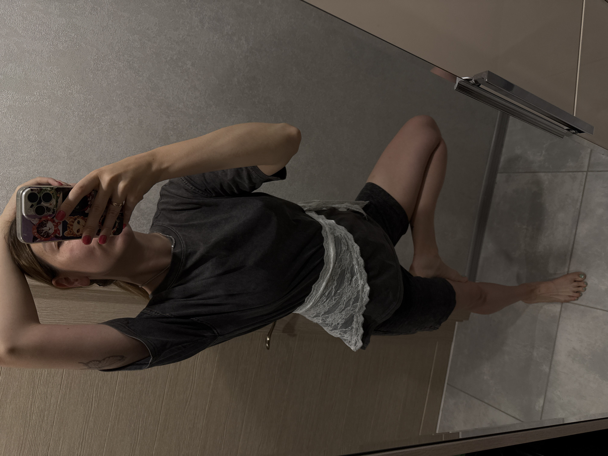 A person is taking a mirror selfie in what appears to be a bathroom. The person is wearing a dark t-shirt, dark shorts, and a white lace sash around their waist. They are standing on one leg, with their other leg bent and raised behind them. The person's left arm is raised above their head, and they are holding a phone in their right hand, which is capturing the reflection. The background shows a tiled floor, a wooden door, and a textured wall. There is also a glimpse of a mirrored surface and a silver handle. The lighting is dim, creating shadows. The scene is indoors and appears to be a private residence. No information about the location being in Moscow, Russia can be determined from the image.