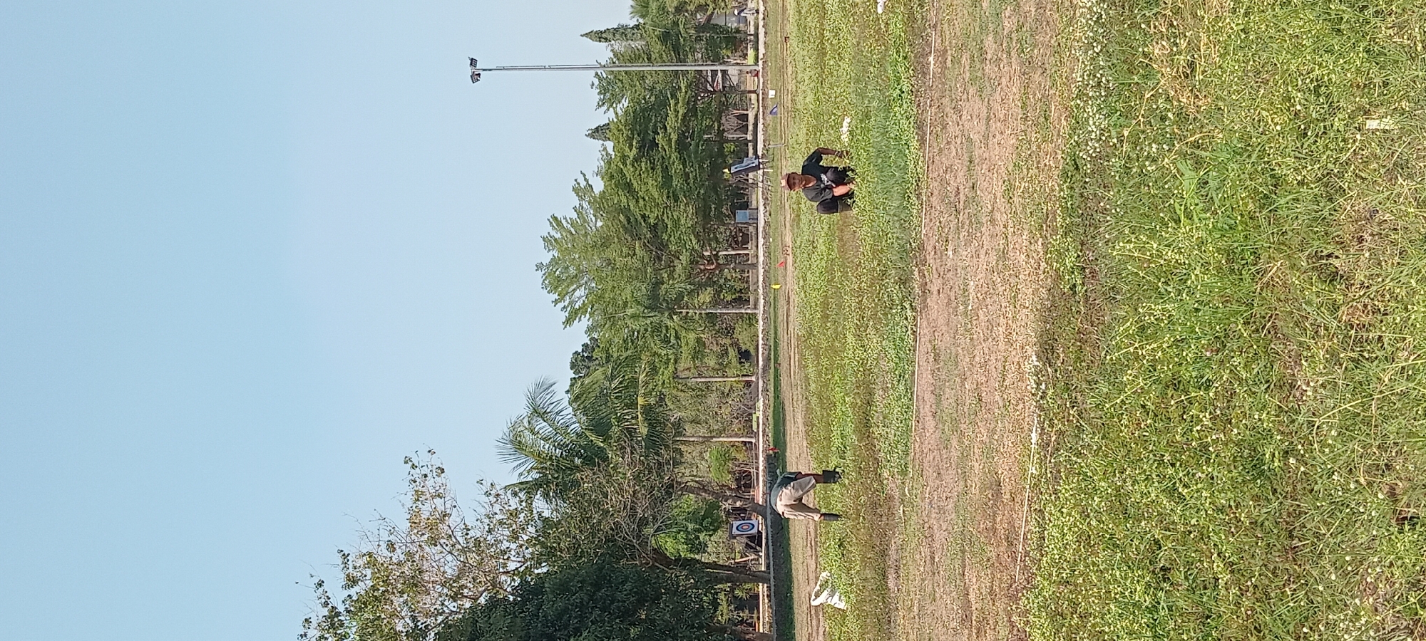In an outdoor scene on a clear day, two individuals are engaged in activities on a grassy field. The field is partially dry and partially covered with green vegetation, with some scattered white flowers. In the background, there is a line of trees, a light pole with two lamps, and what appears to be an archery target.
The first individual, on the left side of the frame and closer to the background, is bending over and appears to be working on the ground. They are wearing a light-colored shirt and darker pants or shorts. A white bag is situated near them.
The second individual, located on the right side of the frame and closer to the foreground, is crouching low to the ground. They are wearing a dark t-shirt and dark pants or shorts.
The overall environment suggests a park or an open recreational area. There is no visual information to confirm the specific location within the city of Tasikmalaya, Indonesia.