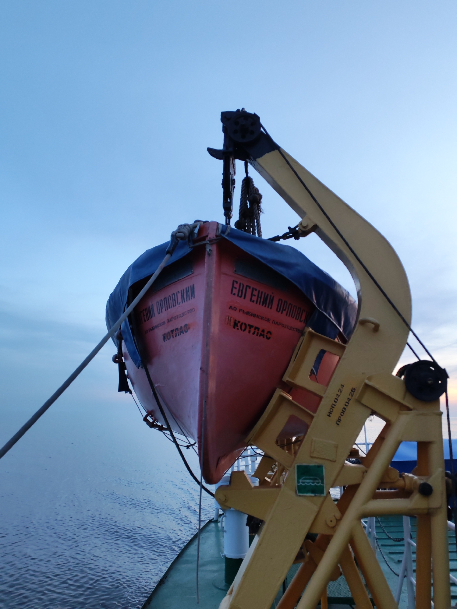 A lifeboat, marked "Евгений Орловский" in Cyrillic and "КОТЛАС" ("Kotlas"), is suspended from a yellow davit system. The boat is predominantly red with black lettering and partially covered by a blue tarp. The davit is mounted on a ship's deck near a body of water under a sky that transitions from blue at the top to a lighter hue near the horizon. Text reading "ИСП.04.24 ПРОВ.04.25." is visible on the davit structure. A green sign with a wave icon is affixed to the davit. The location is indeterminate without further information.