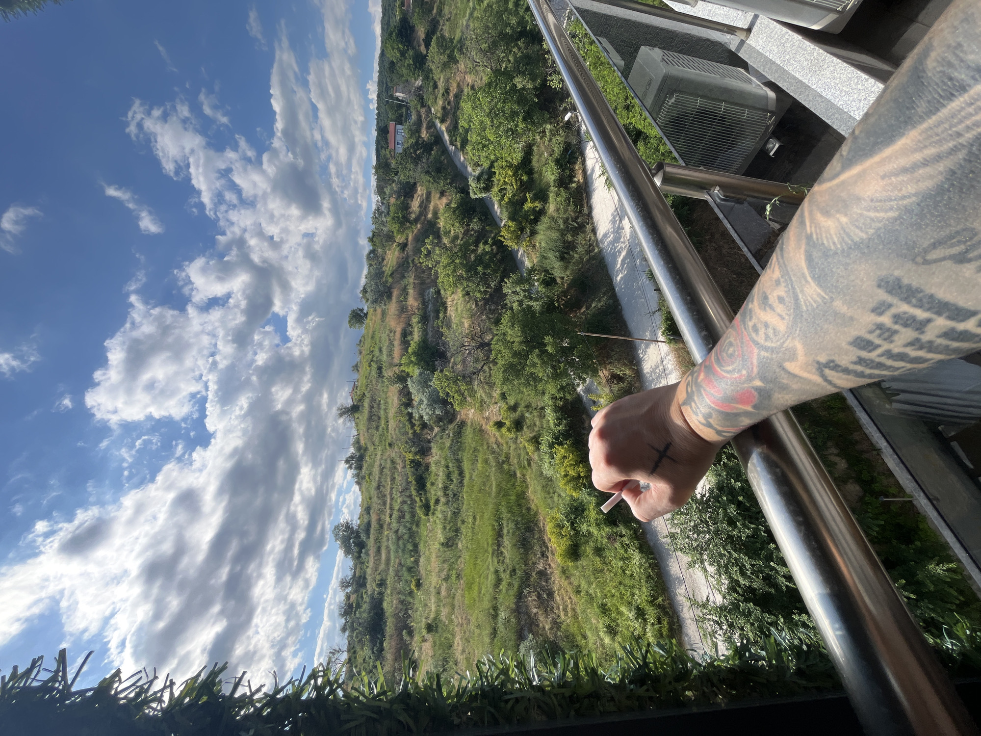 The image shows a first-person perspective of someone holding a cigarette, leaning on a metal railing on a balcony. The balcony overlooks a green, hilly landscape with scattered trees and a winding path. In the background, there are a few houses with red roofs. The sky is blue with scattered white clouds. The person's arm, heavily tattooed, is visible in the foreground, as well as a tattoo on their hand resembling a cross. An air conditioning unit is visible on the balcony to the right. The scene suggests a residential area in Varna, Bulgaria, with a view of the natural surroundings.