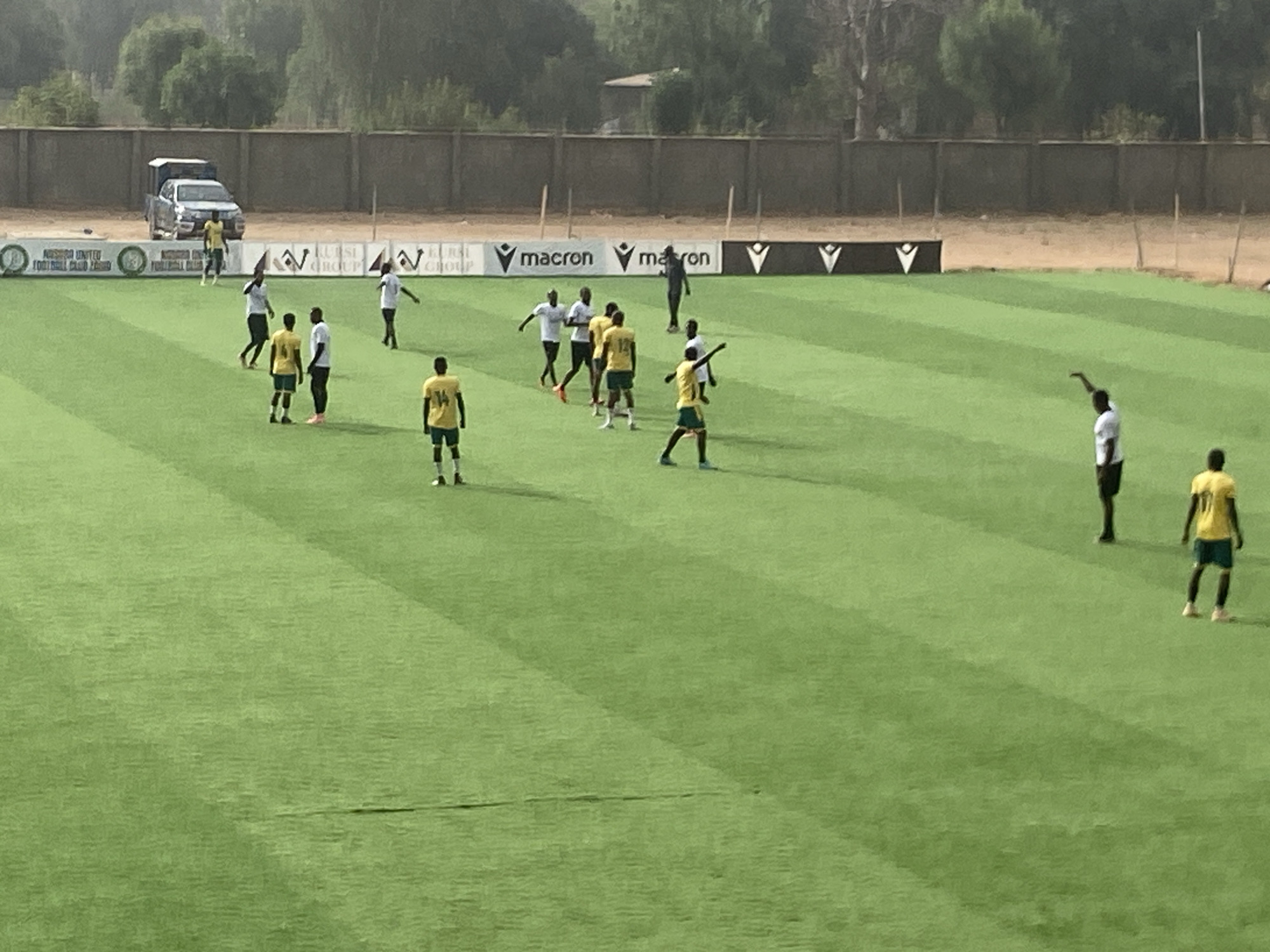 The image depicts a group of people on a green, striped soccer field in Tudun Wada, Nigeria. Most of the individuals are wearing yellow and green soccer uniforms, while others are in white shirts and dark pants. They appear to be stretching or warming up. In the background, a wall with sponsor banners is visible, including brands like "Macron" and "Kurfi Group." A light-colored pickup truck is parked near the wall. Trees are visible beyond the wall. The overall environment suggests a sunny day at a soccer training facility or stadium.