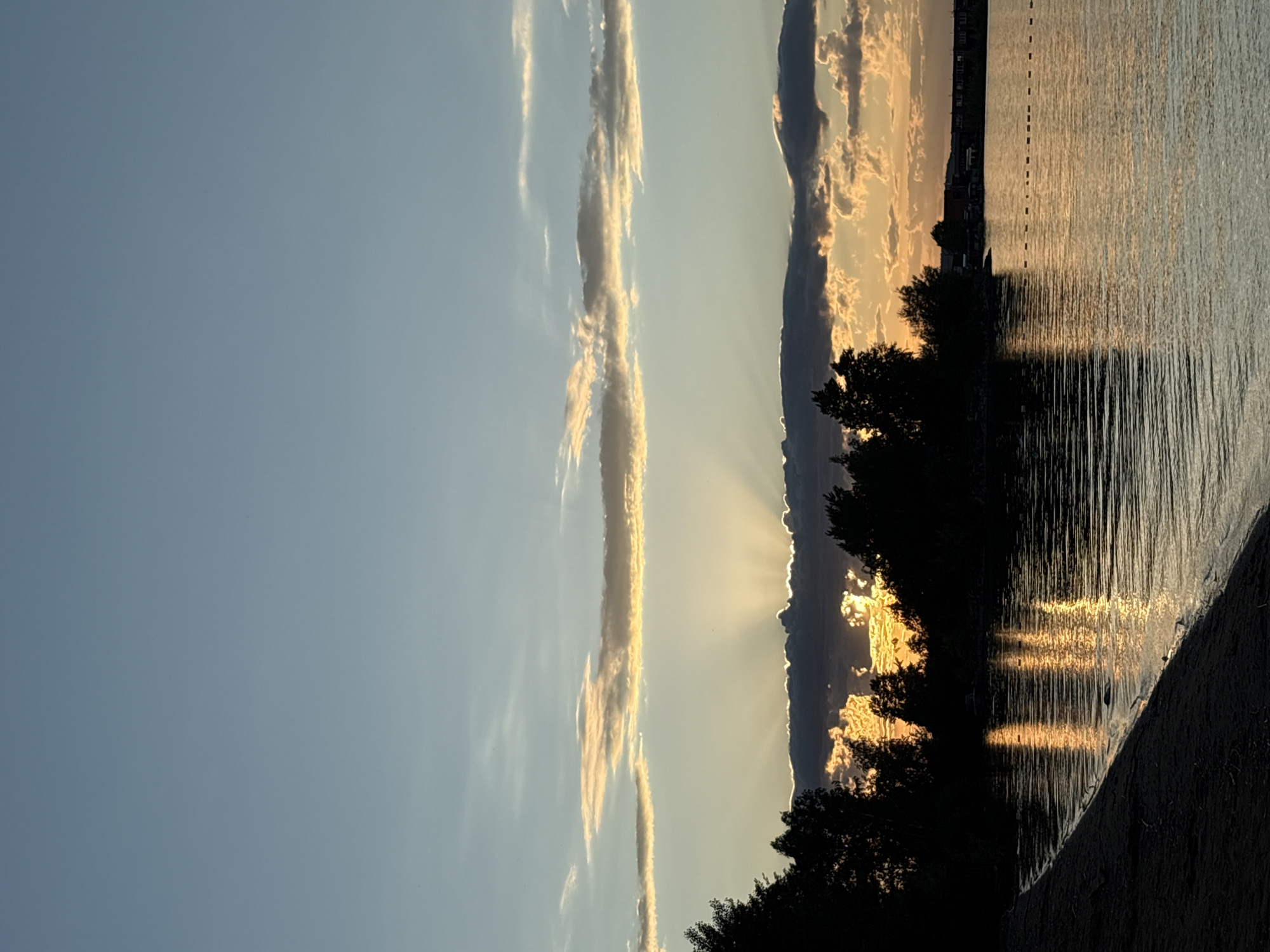 The image, presented in a portrait orientation, captures a serene sunset or sunrise over a large body of water, likely a lake or wide river, in Chaikovsky, Russia. The actual horizon runs vertically through the middle of the frame, separating the expansive sky on the left from the reflective water on the right.
The left side of the image showcases the sky, which transitions from a clear, pale blue at the far left to a warm, golden hue closer to the center, indicating the sun's position. Several long, thin cloud formations are visible, stretching vertically and catching the golden light. Dramatic crepuscular rays fan out from behind a distant landmass or dense cloud bank along the vertical horizon, creating bright streaks across the sky and clouds.
The right side is dominated by the calm surface of the water, which vividly reflects the sun's warm light. These reflections appear as prominent, shimmering vertical streaks of bright orange and yellow, especially in the lower right. Dark silhouettes of trees and foliage line the immediate shoreline at the bottom of the image, providing a stark contrast to the brilliant light on the water. In the distance, along the upper right part of the vertical horizon, faint outlines of buildings or structures are discernible on the far bank. Subtle dashed lines, possibly indicating buoys or markers, can be seen further out on the water's surface. The scene evokes a peaceful and atmospheric late afternoon or early morning ambiance. No visible text is present.