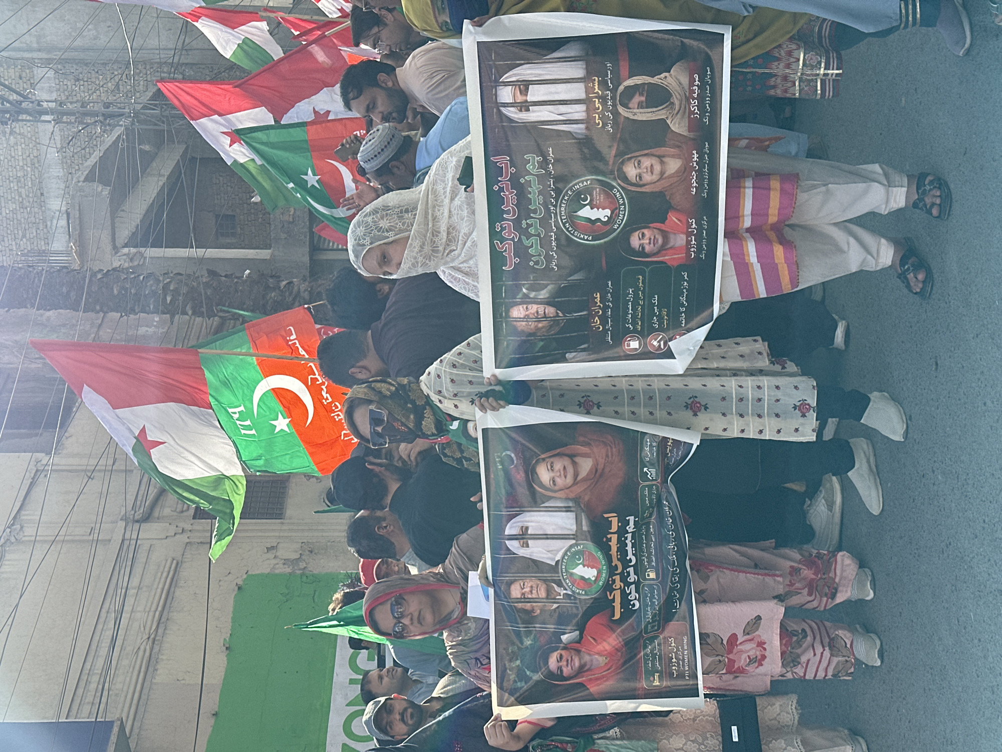 A political demonstration is occurring on a street in Quetta City Tehsil, Pakistan. The crowd is holding flags and signs. The flags are green, white, and orange. The signs have images of women and Imran Khan, with Urdu script. The buildings in the background appear to be multi-storied.