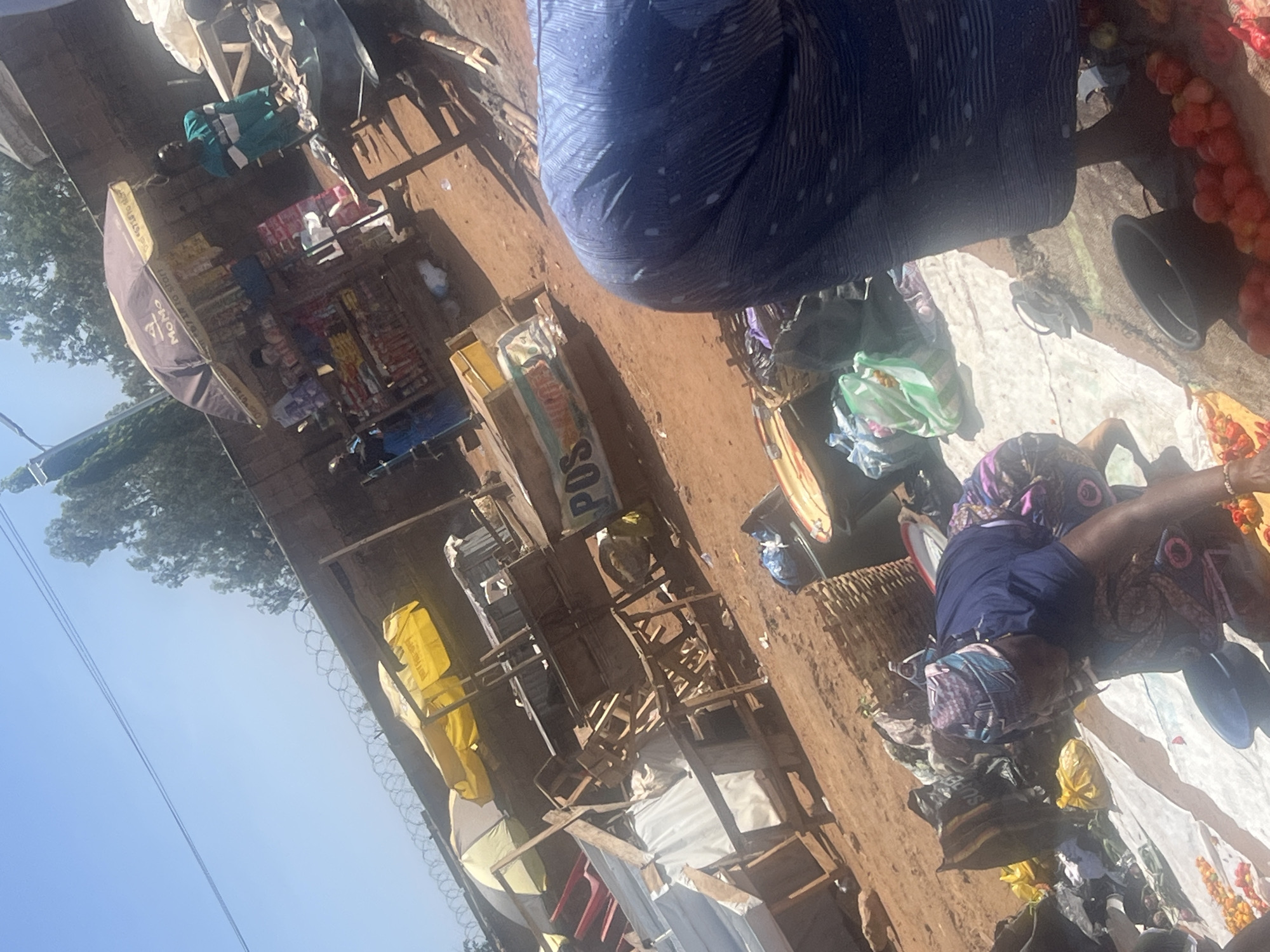 The image shows a street scene in Gura Top, Nigeria, on a sunny day. The ground is reddish-brown. In the foreground, there are people selling items on a white cloth. One person is wearing a blue patterned dress and a patterned headscarf. Beside them are fruits, possibly tomatoes, displayed near a black basin.
In the background, stalls with umbrellas are lined up along the street. One stall has a "MOMO" branded umbrella and displays various goods. Another stall has a "POS" sign. Barbed wire runs along the top of a wall in the mid-ground. The sky is blue with trees visible.