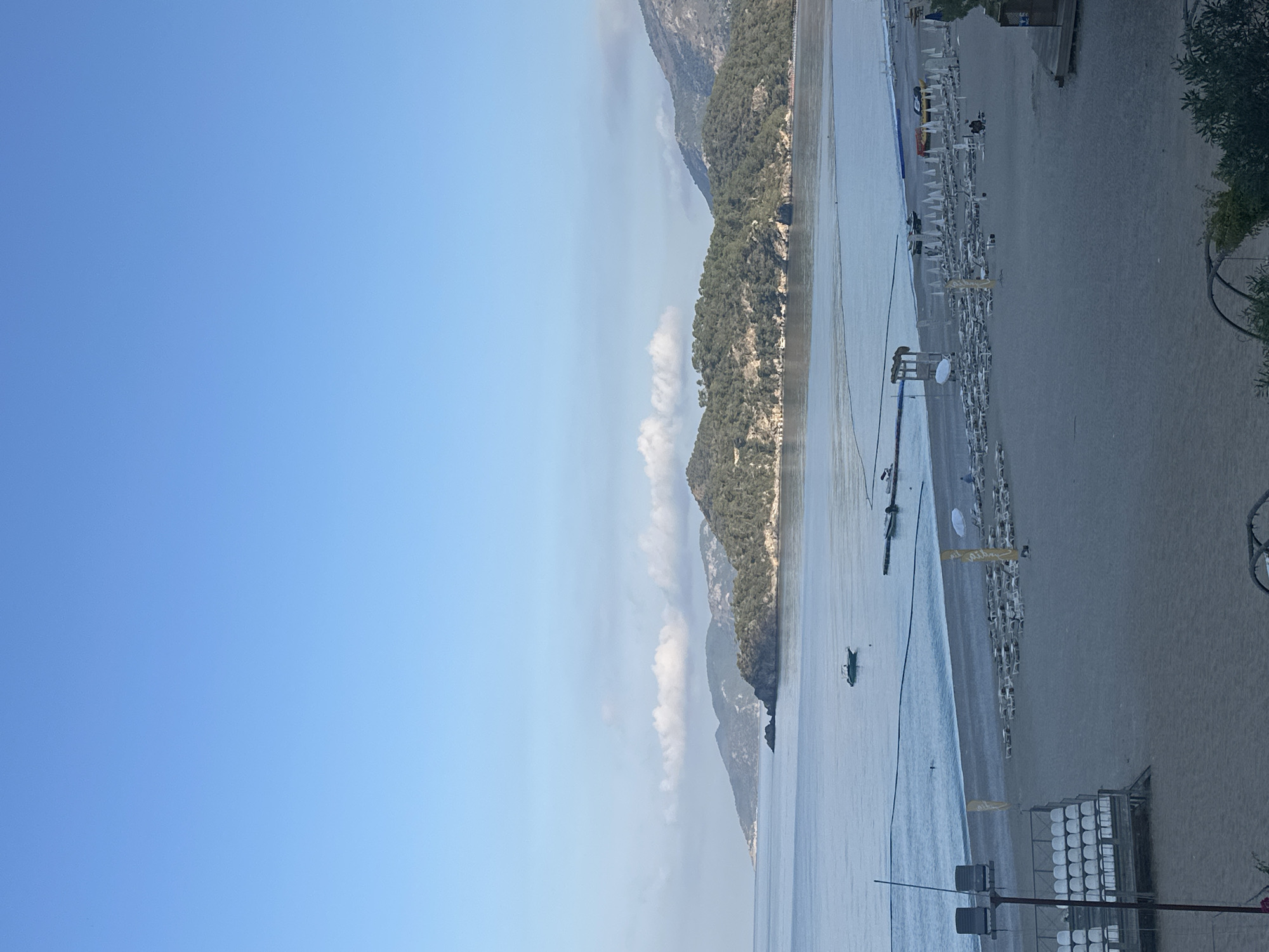 The image captures a wide coastal scene in Fethiye, Turkey, featuring a bay, beach, and mountains under a clear blue sky. The sky, occupying the upper portion, is largely unclouded, with a band of sparse white cumulus clouds extending horizontally across the mid-distance mountains. The water of the bay is calm and appears blue-grey. A small motorboat is anchored in the left-center of the bay. A floating barrier or rope delineates a swimming area closer to the shore, where one individual is visible in the water. A small, dark-colored floating dock is positioned further right.
The coastline is characterized by a series of forested hills and mountains, receding into the distance. The foreground features a wide, light-colored sandy beach. Multiple rows of unoccupied white sun loungers, each accompanied by a closed white umbrella, are neatly arranged across the sand. On the far left of the beach, a multi-tiered seating structure with white seats is prominently displayed. Several yellow flags are interspersed among the sun loungers. A lifeguard tower is visible towards the right side of the beach. A few individuals are present on the beach, some near the sun loungers. A dark utility pole with lights is partially visible in the extreme left foreground, and green foliage occupies the bottom edge of the image.