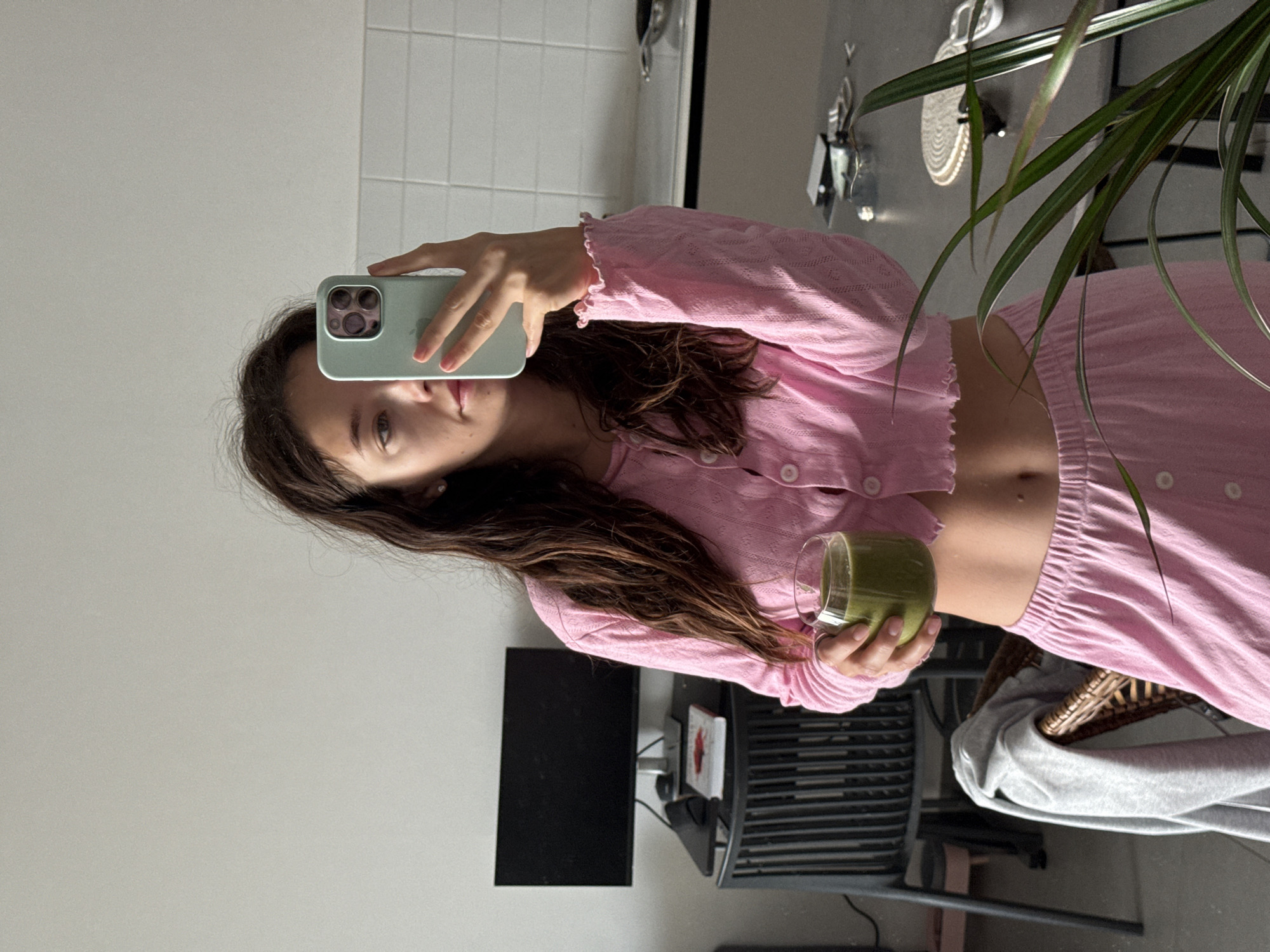 A young woman is taking a selfie in a mirror while holding a green smoothie in a clear glass. She's wearing a pink cardigan with matching pants, revealing her midriff. Her hair is long and brown.
The background shows a partially visible kitchen or bathroom with white tiled walls. Various objects are visible in the reflection including a plant, some decor, and a computer monitor on a desk.
There is no indication from the image that this is in Tel-Aviv, Israel.