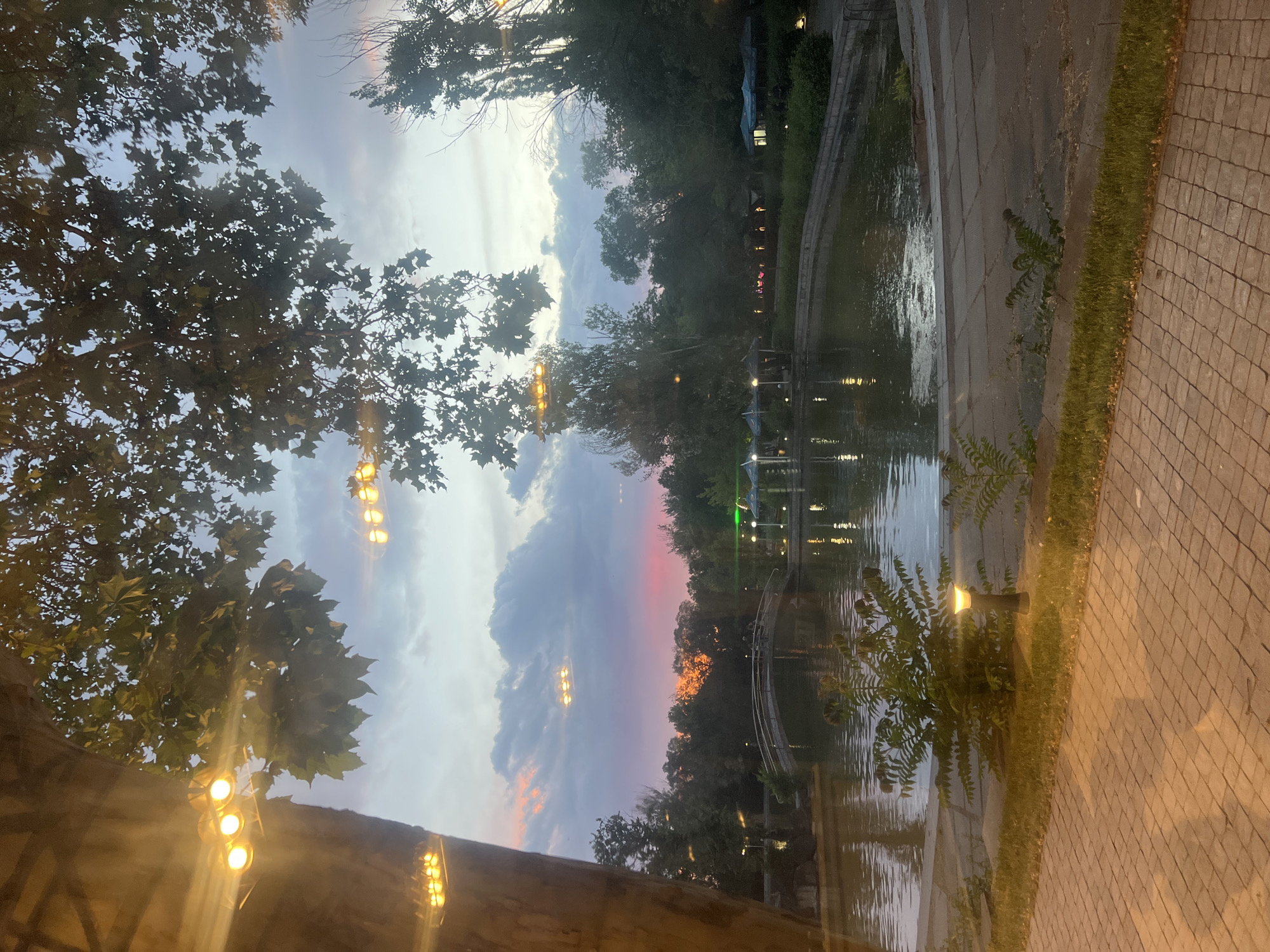 The image depicts a sunset scene at a park. A calm body of water is in the foreground, with a paved walkway to its right and grass to its left. In the distance, a bridge spans the water, and several trees line the park's edge. The sky is a blend of soft blues, purples, and oranges from the setting sun, with scattered clouds. String lights are visible in the trees, and pathway lights illuminate the ground. There are a few people visible on the bridge and near some covered seating areas along the water. The overall atmosphere is peaceful and serene. This scene is likely located in Victory Park in Yerevan, Armenia, known for its lake, bridge, and recreational areas that often feature evening lighting.