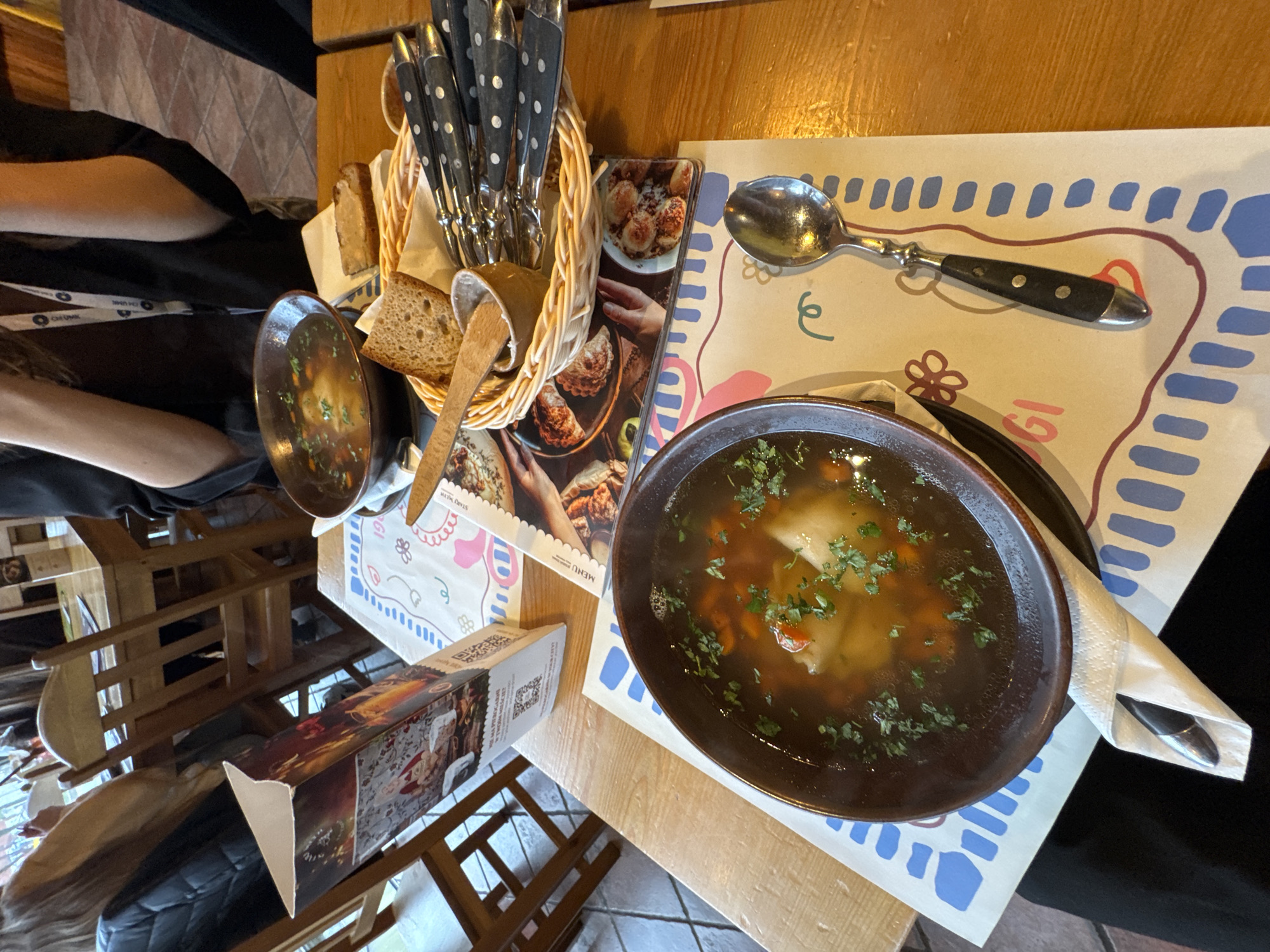 The image depicts a meal at a restaurant, likely in Bydgoszcz, Poland. A table setting includes two bowls of soup (possibly broth-based, with vegetables and garnished with herbs) on patterned placemats. A basket holds bread and silverware, accompanied by a menu or promotional material featuring images of food. A spoon rests on one of the placemats. A person wearing black clothing and a lanyard is seated at the table. The image suggests a dining experience.