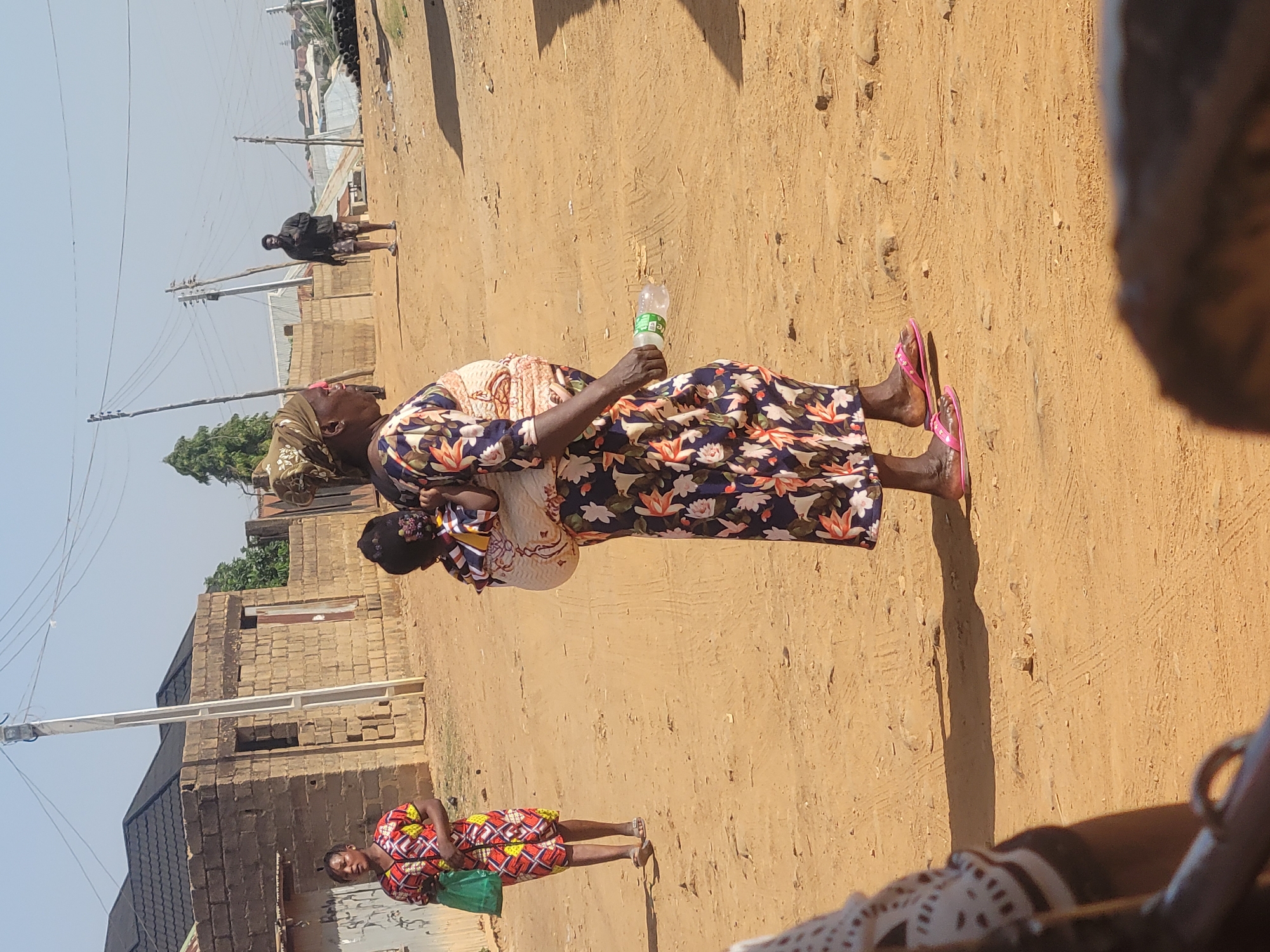 The image depicts an outdoor scene in a residential area of Jos, Nigeria. A woman wearing a floral-patterned dress and a headscarf is standing on a dirt ground, holding a baby on her back. She is also holding a water bottle. Another woman is standing nearby, wearing a patterned dress and holding a green bag. In the background, there is a brick building and electrical poles. A person is walking further down the road, and there is some other indiscernible activity in the distance. The ground appears to be sandy.