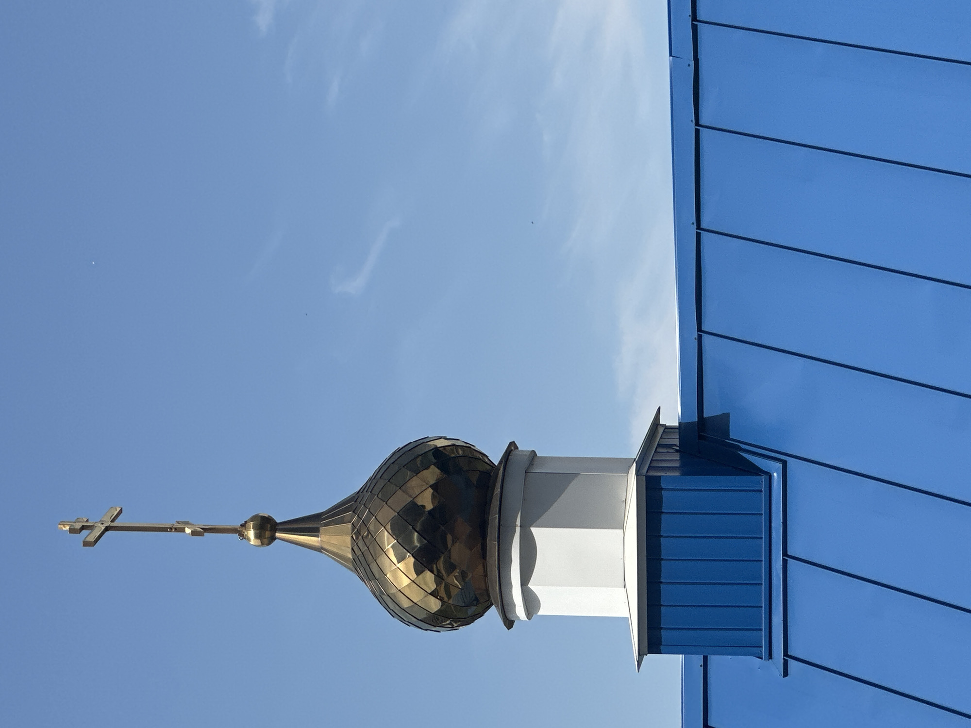 A low-angle shot shows the golden dome and cross of a church against a clear blue sky. The roof of the church is clad in blue metal siding. The location is likely Novosibirsk, Russia. The scene is focused on the architectural details of the church against a bright, uncluttered sky.
