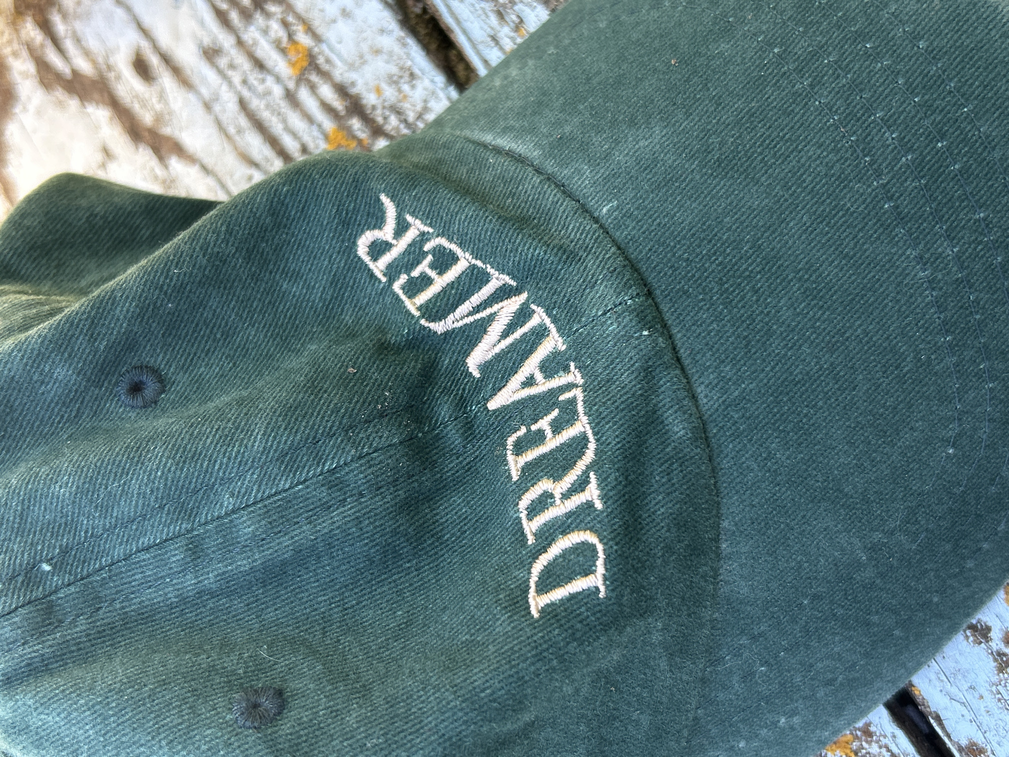 The image shows a close-up view of a dark green baseball cap. The word "DREAMER" is embroidered in white on the side of the cap, with the letters curved to follow the shape of the cap. The fabric of the cap has a textured weave. The cap is resting on a surface that appears to be weathered wooden planks, with some lighter colored paint or stain and yellow splotches visible. The lighting suggests it is daytime, with shadows indicating the light source is coming from the side. The overall setting appears to be outdoors, possibly in a rustic or casual environment, with no specific identifiable landmarks within the frame. The image is a static, close-up shot of the cap, with no activity or event depicted beyond the object itself.