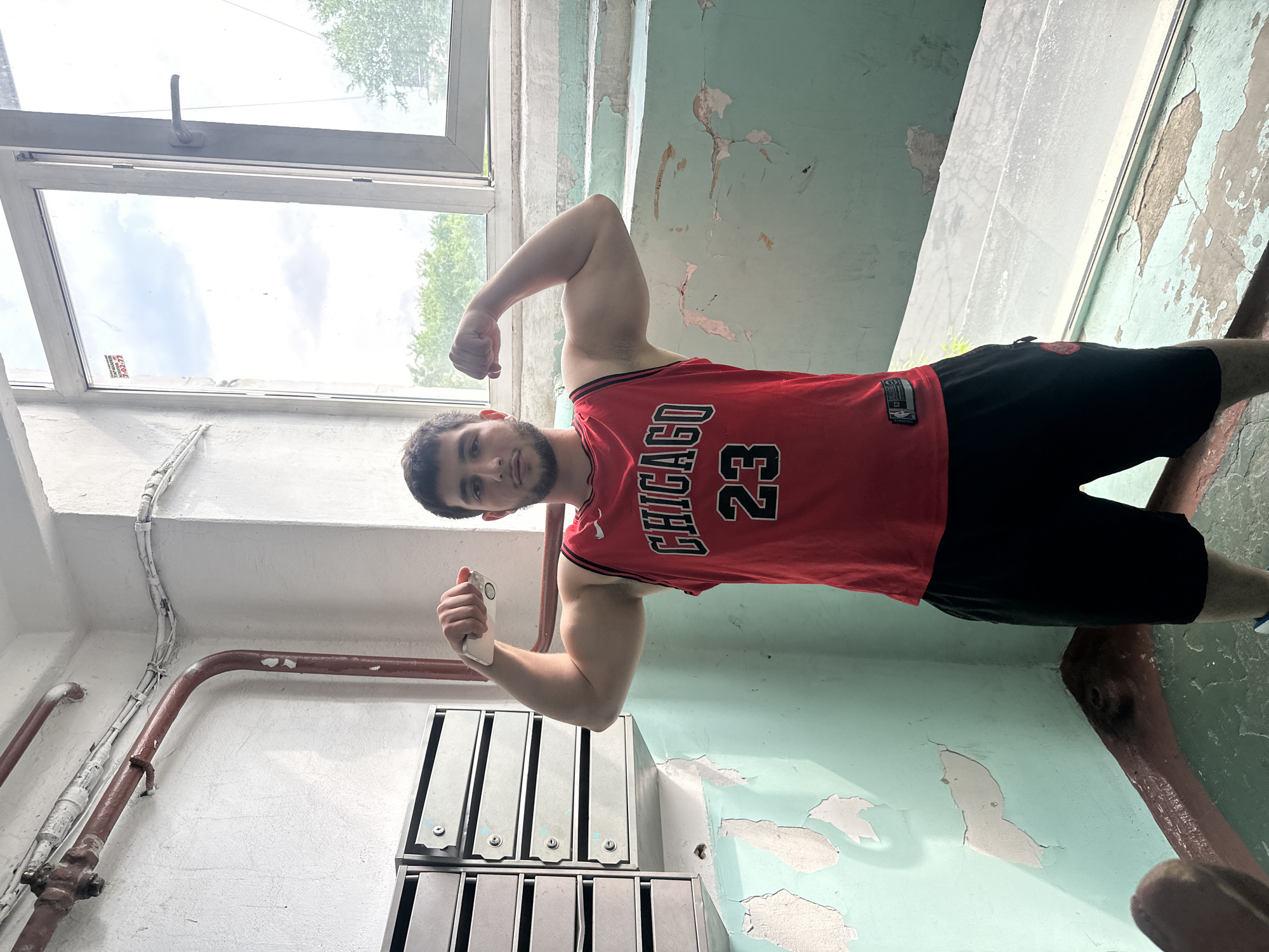 An adult male with dark hair and a beard, wearing a red Chicago Bulls basketball jersey with the number 23 and black shorts, stands in a stairwell. He is flexing his biceps, holding a white smartphone in his right hand. The stairwell is located in a building in Birobidzhan, Russia. The walls are painted a peeling light green, and there are metal mailboxes mounted on the left wall. A large window is visible in the background, showing trees and a cloudy sky. Pipes and electrical conduits are visible along the ceiling and wall. The overall environment appears worn and somewhat neglected.