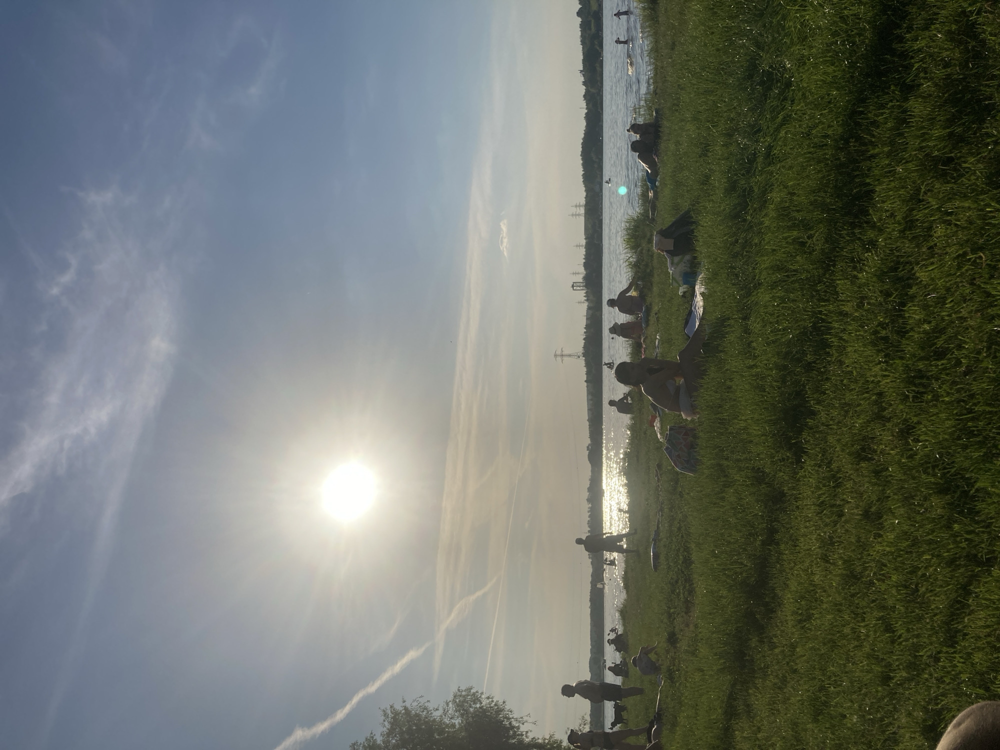 The image captures a vibrant, sun-drenched late afternoon or early evening scene at a lakeside or riverside location in Kapustino, Russia. The bright, low-hanging sun, positioned in the upper-left, creates a strong lens flare and backlights the entire scene, casting most foreground and midground subjects into silhouette. The sky is a clear light blue with delicate wispy clouds and prominent, crossing aircraft contrails.
A large body of water, shimmering with reflected sunlight, stretches across the middle ground. On the far shore, a dense line of trees is visible, punctuated by several large power line pylons and industrial structures, indicating a mixed natural and developed landscape.
The immediate foreground consists of lush, green grass, illuminated by the low sun, with subtle shadows extending towards the viewer. Numerous people are scattered across the grassy bank and in the water. On the left side of the bank, several individuals are standing, with one person appearing to interact with a dog nearby. Throughout the midground, various figures are seen relaxing on the grass, some lying down as if sunbathing, others sitting upright on towels or blankets. Several people are partially or fully submerged in the water, with ripples on the surface indicating movement. The overall impression is one of leisure and enjoyment of the water and sunshine.