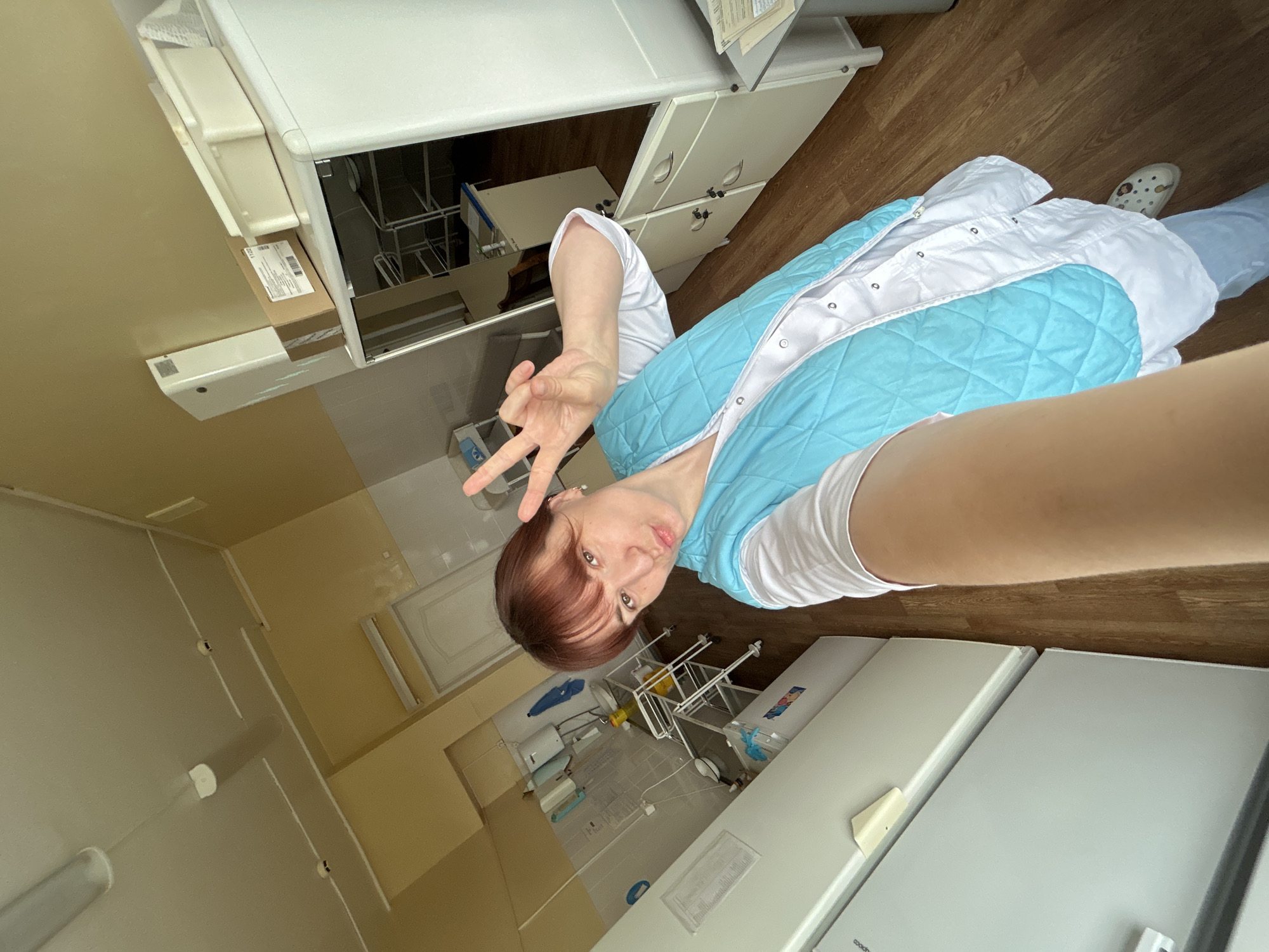 A woman poses for a selfie, giving a peace sign, in what appears to be a clinical or laboratory setting in Фоки, Russia. She looks directly at the camera with a slight smile. She has reddish-brown hair with bangs and is wearing a white long-sleeved shirt underneath a light blue quilted vest or jacket with white trim and snap buttons. Light blue pants and a light-colored Croc shoe on her visible right foot complete her attire.
The room features a wood-patterned laminate floor and light-colored, possibly beige or cream, walls. The background is filled with functional white and light-colored furniture. To the left of the woman, white cabinets, some with drawers, are visible. One cabinet door is open, revealing shelves inside. A brown cardboard box with a barcode and the numbers "1122" is placed on a shelf, near a white wall-mounted device. Further into the background, there's what looks like a sink area, a metal rack or trolley, and two large white upright appliances, which appear to be refrigerators or freezers. The brand "coolpad" is partially visible on one of these appliances. Linear fluorescent lighting fixtures are present on the ceiling and walls. The overall environment suggests a professional and clean workspace.