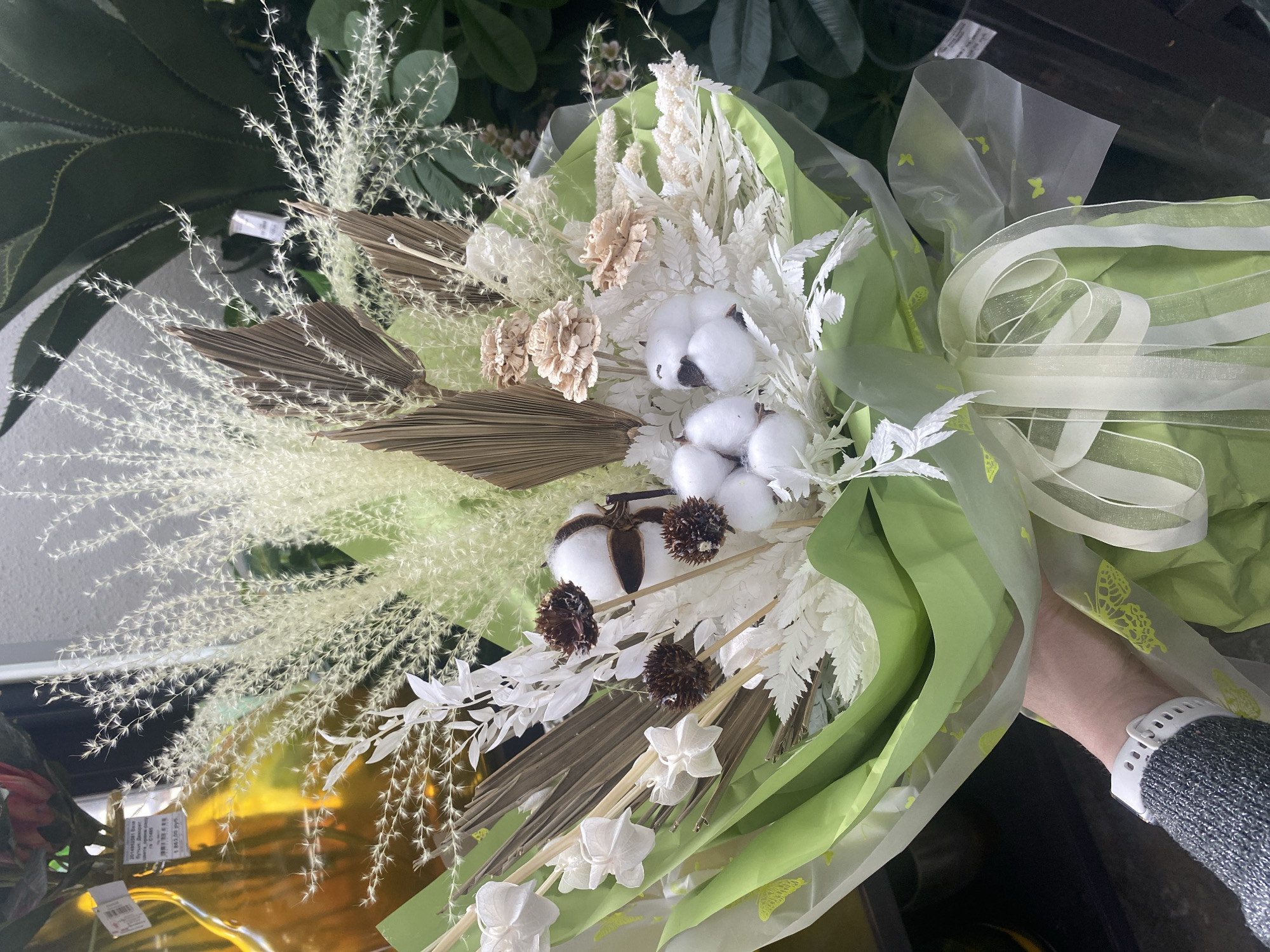 The image depicts a floral arrangement held in someone's hand. The bouquet consists of dried flowers and foliage in shades of white, beige, and brown. Elements include feathery sprays, dried palm leaves, cotton bolls, and paper flowers. The arrangement is wrapped in light green and transparent cellophane with butterfly motifs and tied with a sheer ribbon with white stripes.
While the prompt requests the location to be identified as Yekaterinburg, Russia, there is no visual information in the image itself to support this claim. The image does not contain any recognizable landmarks, signs in Cyrillic script, or other elements that would definitively place it in that city.