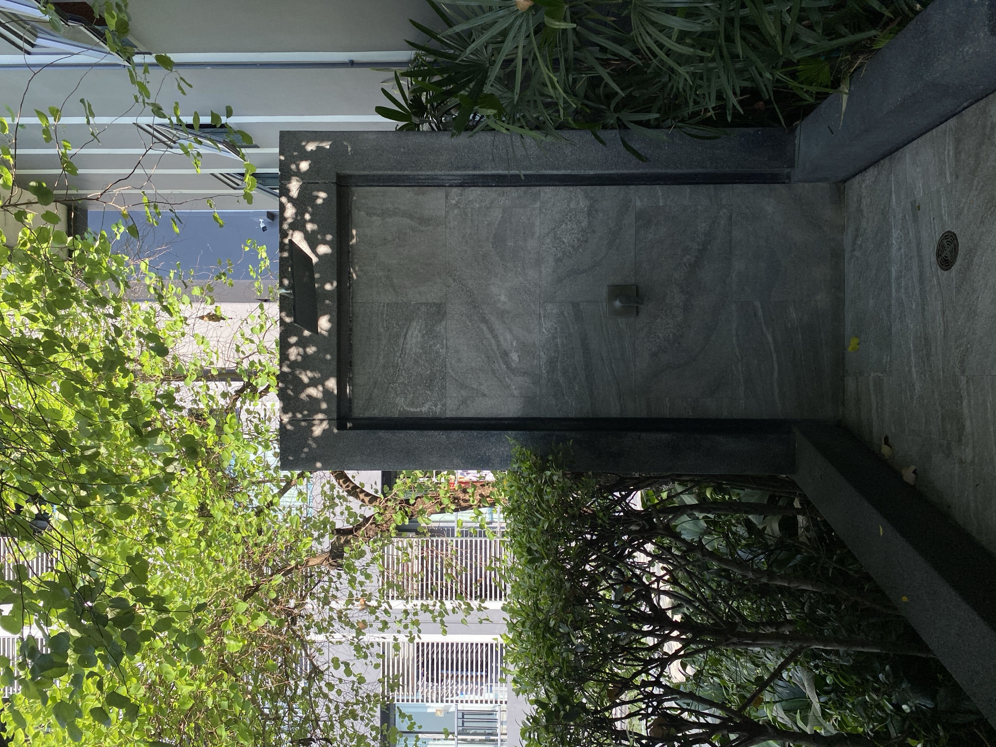 The image captures an outdoor space, likely in an urban setting, possibly Bangkok, Thailand given the vegetation. A gray tile wall with a mounted handle is the central structure. Lush greenery, including what appear to be various types of leafy plants and trees, surrounds the wall, creating a natural barrier. The wall is bordered by a dark, possibly granite frame. To the right of the wall, the ground is paved with gray tiles and features a drain. The sunlight is diffused, creating soft shadows. Portions of a building with white vertical bars are seen through the foliage, suggesting residential or commercial architecture.