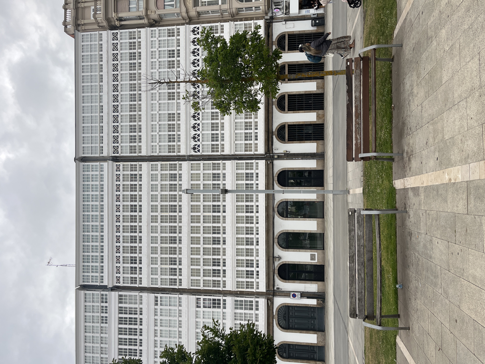 This is an exterior shot of a building in A Coruña, Spain. The building is a large, white structure with numerous windows. The facade is characterized by rows of white-framed windows and decorative elements. At ground level, the building has arched doorways and ground-level openings with dark ironwork. A tree partially obscures the view of the building.
In the foreground, there are benches on a paved sidewalk, with a narrow strip of grass between the sidewalk and the building. A person is sitting on one of the benches. The sky is overcast. A light post is between the building and the benches.