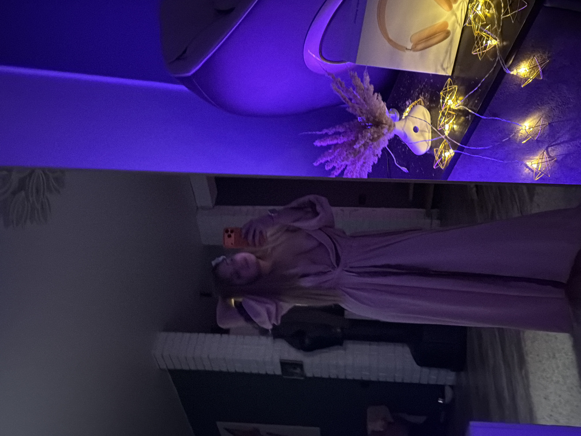 A woman in a long-sleeved, light pink dress is taking a selfie with a phone. She has light-colored hair and is standing indoors. Behind her is a white brick fireplace with dark items placed on it. Above, there's a shelf or table with decorative items, including a white vase with pampas grass, headphones, and star-shaped string lights. The top portion of the frame is illuminated with a blue light. The location is possibly Большая Шильна, Russia, based on the user's prompt.