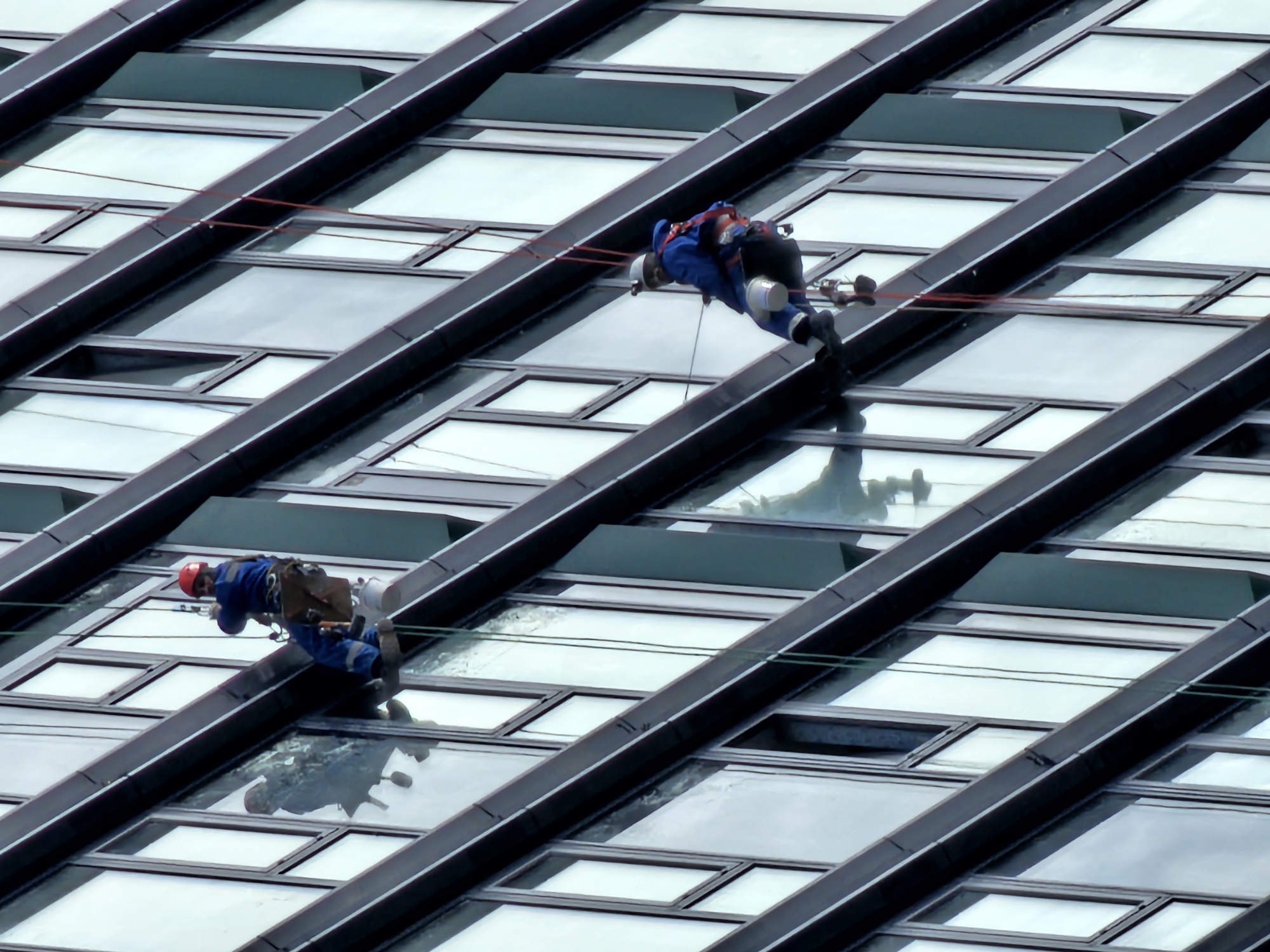 Two window washers suspended on ropes are cleaning windows on the exterior of a modern building. The building features a grid of large rectangular windows framed by dark horizontal and diagonal beams. The workers are wearing blue jumpsuits and safety helmets. One worker has a red helmet. The windows reflect the sky, and there are reflections of the workers on the glass surfaces. Moscow, Russia.