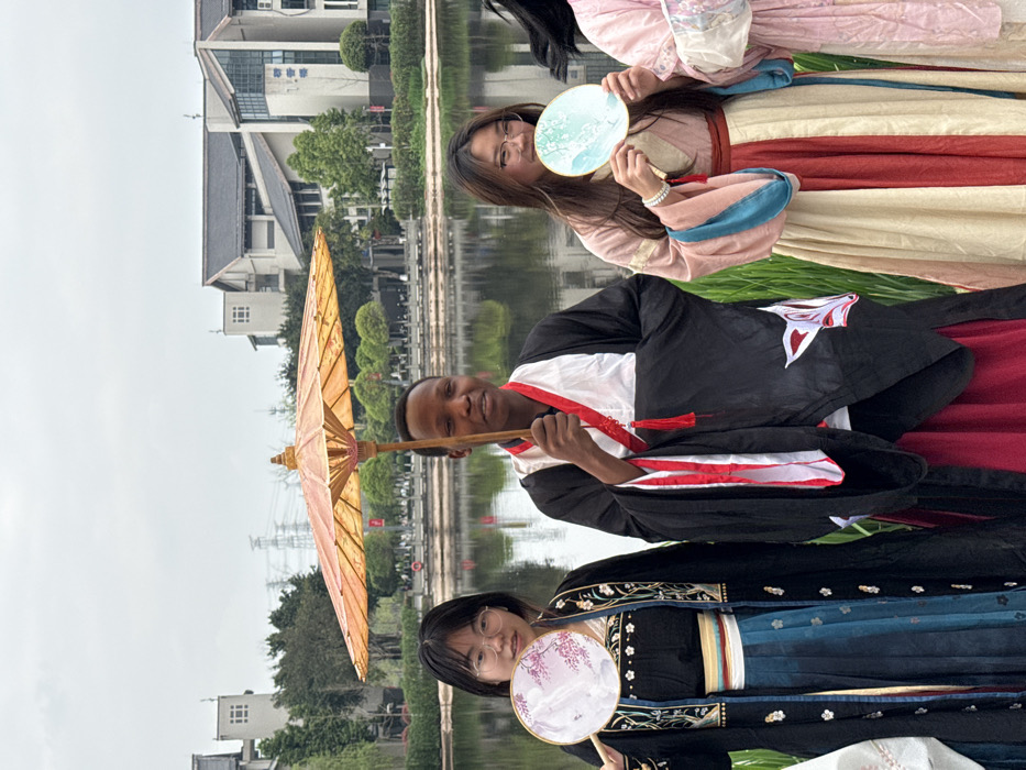 This image captures five individuals, three fully visible, posing outdoors beside a calm body of water in Jiangjin, China. The central figure is a Black male holding an open orange oil-paper umbrella overhead, wearing a black and white traditional Hanfu with red trim and tassels. To his left, an East Asian female in eyeglasses holds a round fan adorned with a pink floral design, dressed in a black and blue Hanfu featuring embroidered details. To his right, another East Asian female in eyeglasses holds a round fan with a light blue and green nature-themed design, attired in a light pink, cream, and red Hanfu with teal sleeve accents. Two additional individuals are partially visible on the far left and far right, also in Hanfu-style garments. All visible subjects are looking towards the camera.
The immediate foreground features green grass or reeds. The middle ground is occupied by a still body of water, reflecting the overcast sky and distant structures. The background consists of several multi-story buildings with a mix of traditional Chinese rooflines and modern architectural elements, suggesting a university campus or cultural complex. One building on the right bears the Chinese characters "博雅楼" (Bóyǎ Lóu). Lush green trees and shrubs surround the buildings and line the far bank of the water. In the distant background, a large metal power transmission tower and associated lines are visible against the grey, overcast sky.