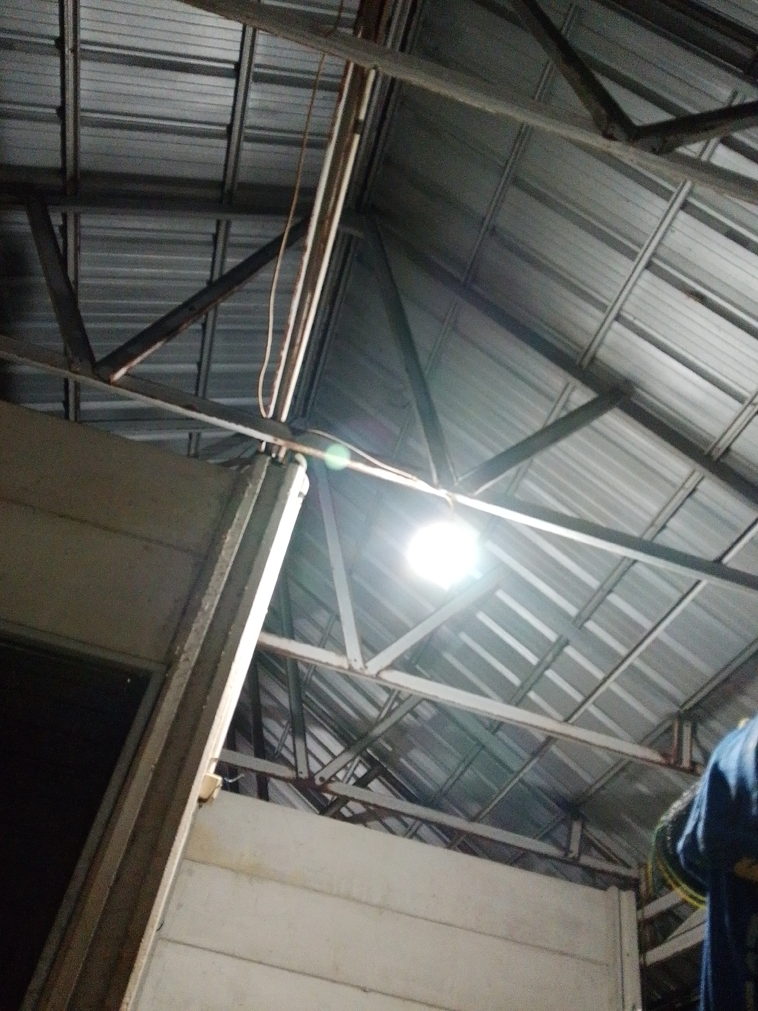 The image shows the interior of a building, possibly a warehouse or industrial structure, in Bukit Ketok, Indonesia. The roof is corrugated metal supported by a framework of steel beams and struts. A bright light is suspended from the ceiling. A partial view of a white wall and a dark doorway are visible on the left. A person wearing a blue shirt appears on the right edge of the frame.