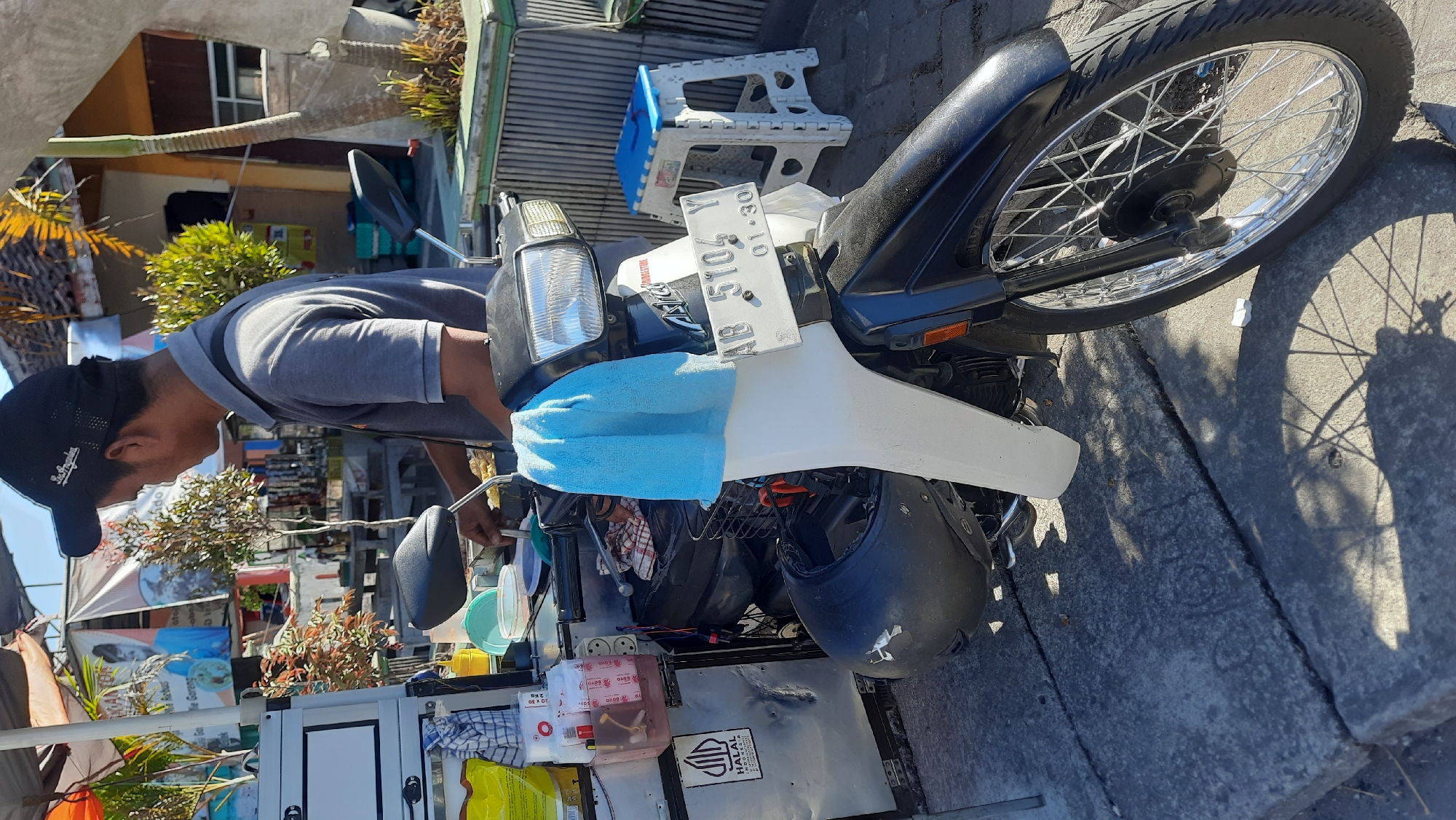 The image shows a street vendor in Nanggulan, Indonesia, preparing food. The vendor, a man wearing a gray polo shirt and a black baseball cap, is standing behind a mobile food cart attached to a motorcycle. The cart is equipped with various containers and ingredients, including sauces and packaged food items. A blue cloth is draped over the front of the motorcycle. In the background, there are banners with text in Indonesian, a palm tree, and a white plastic stool. The scene appears to be outdoors, with sunlight casting shadows on the concrete ground. The motorcycle's license plate reads "AB 5104 Y".