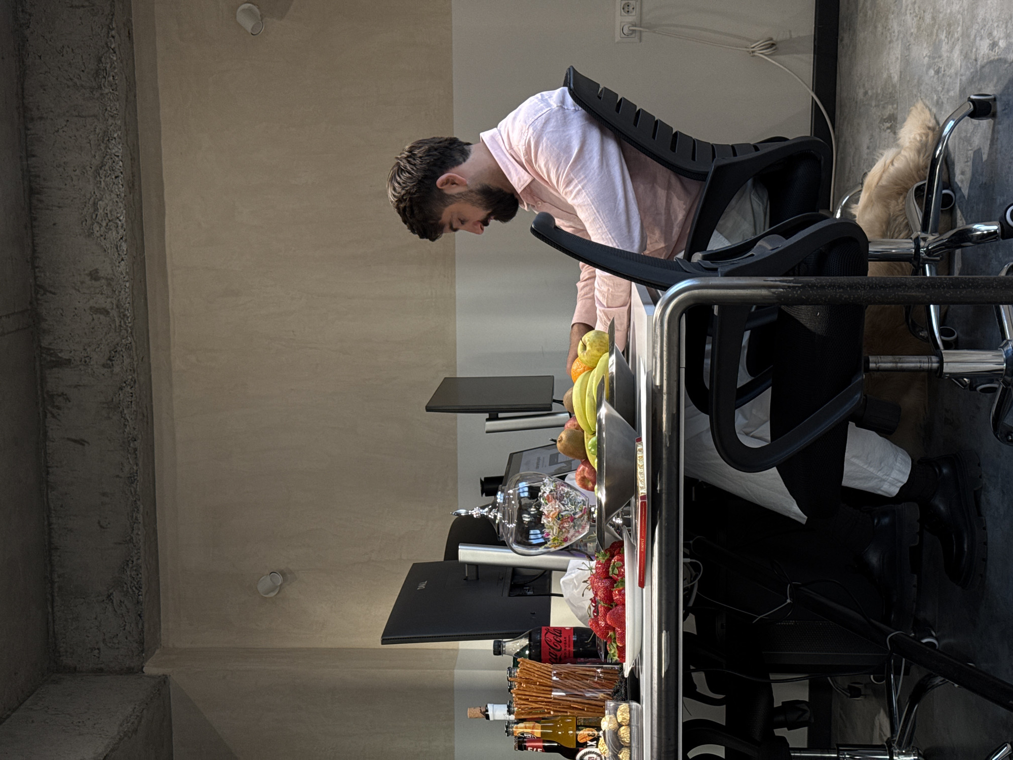 An adult male, approximately 25-35 years old with dark hair and a beard, is seated in a black ergonomic office chair at a desk in an indoor office environment in Yerevan, Armenia. He wears a light pink long-sleeved shirt and white pants, with black shoes and socks. He is focused on a task, looking down at the desk where a computer monitor is visible.
The desk, light-colored with metallic legs, is laden with various items: a tray of small pastries, several bottles including red wine, beer, and Coca-Cola Zero Sugar, a tall glass filled with pretzel sticks, a clear glass candy dish with a decorative lid containing colorful candies, a white plate piled with fresh strawberries, a silver bowl holding bananas, apples, and oranges, and a red "Merci" chocolate bar package. Two black computer monitors (one a Dell brand) are positioned on the desk.
The room features a two-tone wall, with a lighter beige/off-white lower section and a darker, textured upper section, possibly exposed concrete. The ceiling is exposed concrete with visible beams. White cylindrical light fixtures are mounted on the upper wall. The floor is light gray concrete. A white Type F power outlet with a plugged-in cable is visible on the right wall. Partially visible under the desk on the right, a light-colored, long-haired dog is resting on the floor.