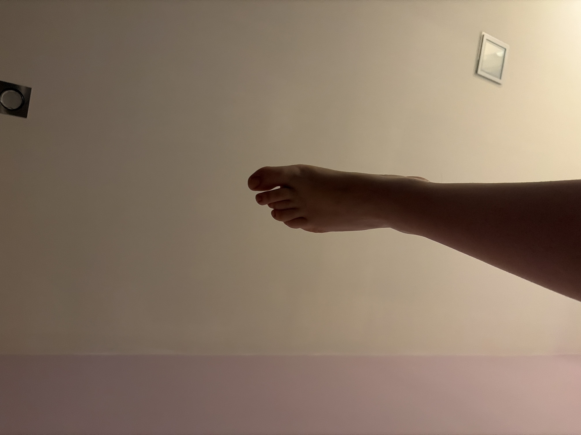 A person's left foot and lower leg are visible, angled upwards against a plain, light-colored wall. The foot is bare and the toes are distinct. The lighting suggests an indoor environment, potentially a room with recessed ceiling lights, one of which is partially visible at the top of the frame, and another square light fixture lower down. The wall on the left appears to be a slightly different shade, possibly a corner where two walls meet or a painted section. There are no other people or objects clearly identifiable in the frame. The image does not contain any information that would allow for a precise location within the city of Dobrich, Bulgaria.