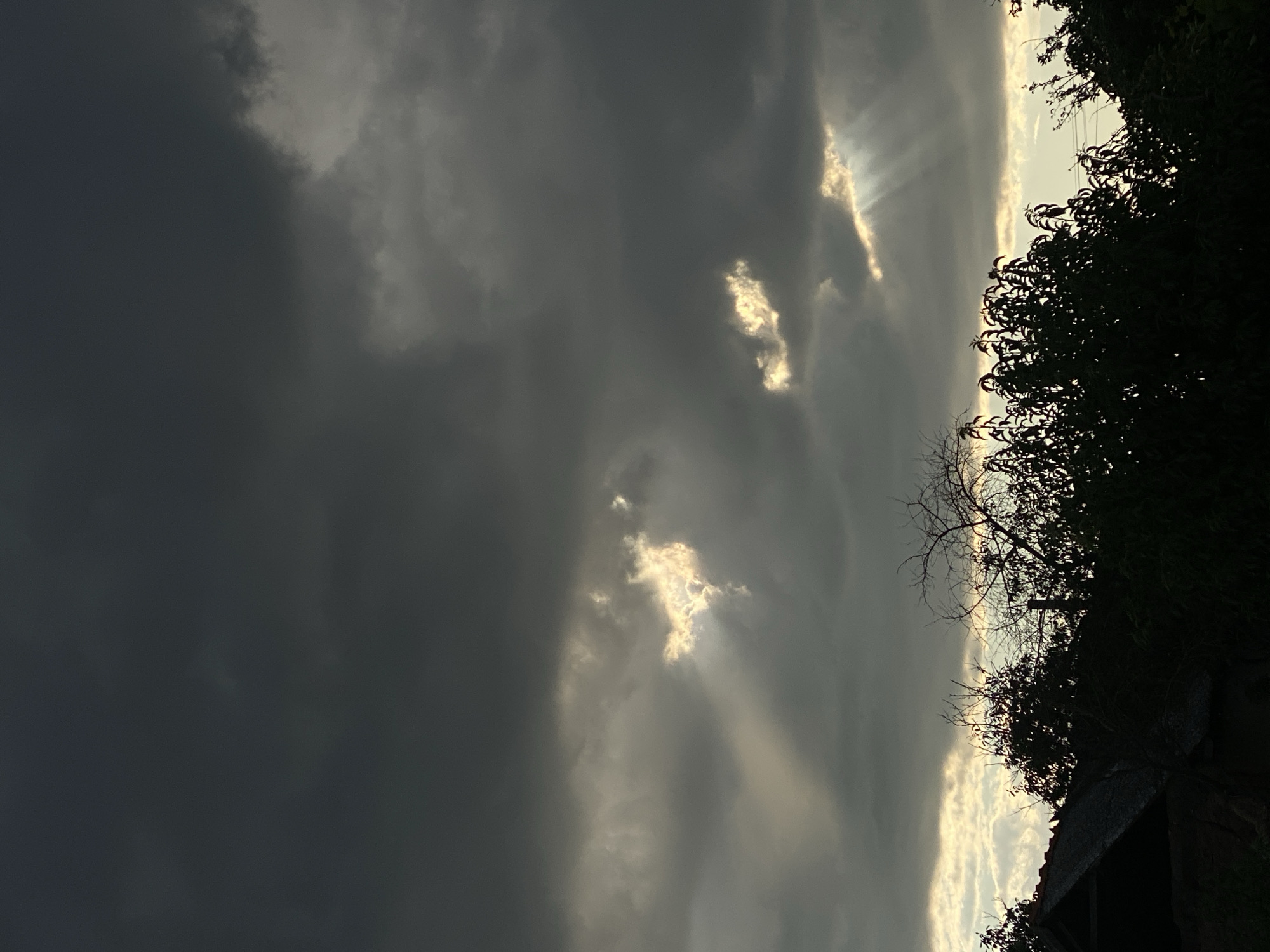 The image depicts a dramatic sky over Cairaclia, Moldova, with heavy, dark clouds obscuring most of the sun. However, several shafts of light pierce through gaps in the clouds, creating visible beams that illuminate portions of the sky. In the foreground, silhouetted trees and foliage are visible, along with the dark roof of a structure that appears to be a house or shed. The overall mood is somber and atmospheric, suggesting impending rain or a storm. There are no people or animals visible in the frame.