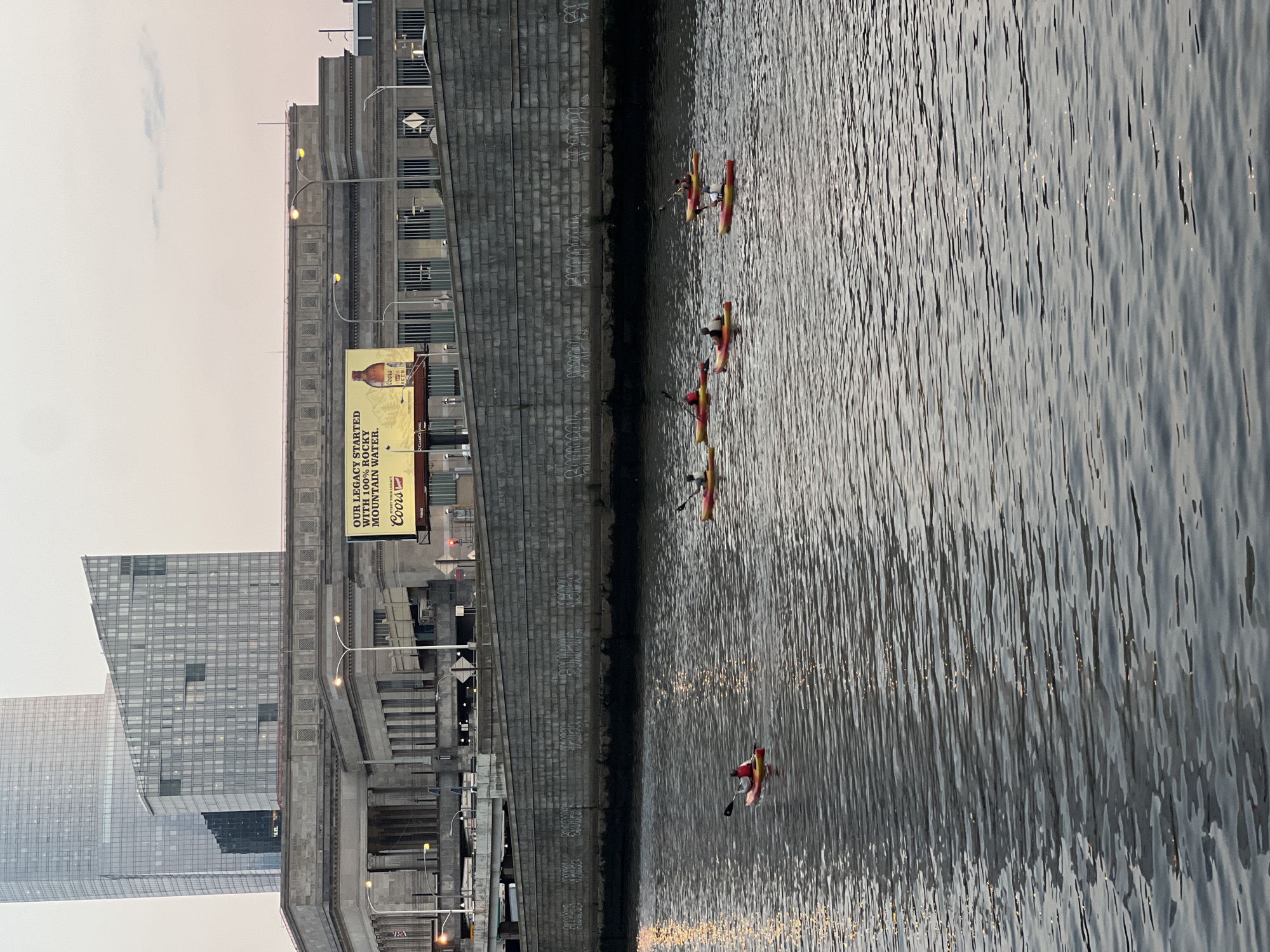 In Philadelphia, Pennsylvania, USA, a group of six kayakers navigate a body of water. They are positioned in the foreground and mid-ground of the image. The kayakers are on individual kayaks and appear to be paddling, suggesting they are engaged in recreational activity. The water reflects the soft light of what appears to be dusk or dawn.
Behind the kayakers, a large, textured stone wall runs horizontally across the mid-ground, separating the water from the elevated structures above. This wall shows signs of graffiti. Above the wall, a long, classical-style building with large windows and columns forms the backdrop. A prominent billboard advertising Coors Light is mounted on this building. Further in the background, modern skyscrapers, one with a distinctive angular design, dominate the skyline, indicating an urban environment. Streetlights are visible along the elevated structures and on the building. The overall scene suggests a metropolitan setting with a blend of historical architecture and modern development.