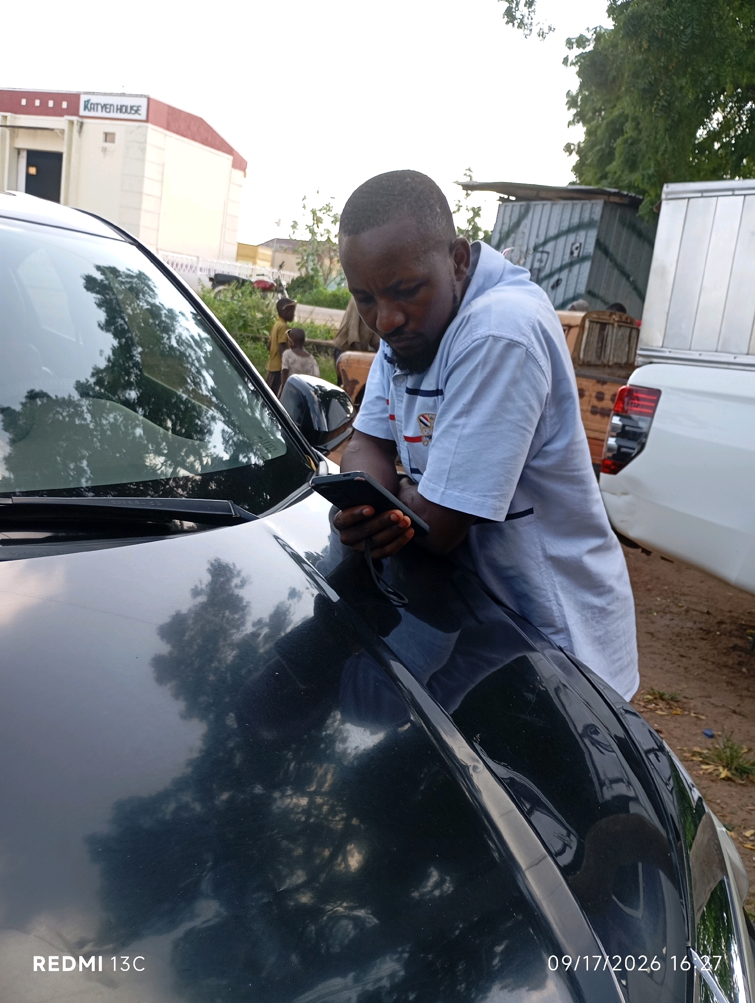 In Shendam, Nigeria, a man with dark skin and a short haircut is leaning on the hood of a black car. He is wearing a light blue collared shirt with dark blue and red trim on the collar and sleeves. He is holding a black smartphone in his hands, looking down at it. Behind the car, a building with the sign "KATYEN HOUSE" is visible. Other vehicles, including a white pickup truck, and several people, including two children, are in the background. The environment appears to be an outdoor area, possibly a street or parking lot, with trees and some scattered foliage. The image is dated 09/17/2026 at 16:27.