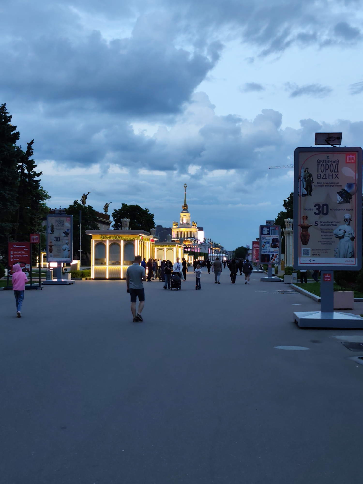 The image captures a view down a wide pedestrian walkway in Moscow, Russia, at dusk or twilight. On either side of the walkway, there are advertising stands with large signs that read "Музейный Город ВДНХ" (Museum City VDNKh) and provide information about the VDNKh. People are walking along the walkway, and a small number of illuminated kiosks or small buildings are visible in the middle ground. The architecture of the buildings and signage are typical of a Soviet-era aesthetic. The sky is cloudy and a deep blue-grey color. In the background, a tower with a spire rises above the other buildings, indicating this is likely the main pavilion or entrance to the VDNKh exhibition center. The walkway is made of asphalt or concrete. A man is walking towards the camera in the foreground, while a person in a pink hoodie is walking away. Other people are scattered along the path, some with strollers. Trees are visible on the left side, adding greenery to the scene.