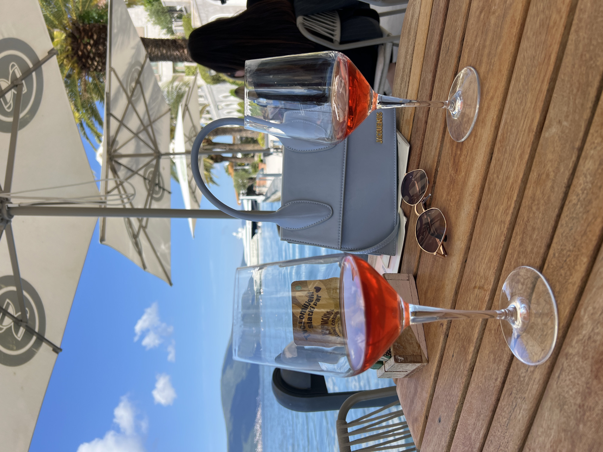 An outdoor cafe scene shows a wooden slatted table set with two glasses of rosé wine, a light blue handbag, and a pair of sunglasses. The image is taken from a slightly elevated perspective, looking towards a body of water with mountains in the distance.
On the table, a light blue (or gray) Jacquemus handbag is prominently placed, with "JACQUEMUS" visible in gold lettering. To its right are a pair of round-framed sunglasses. Two stemmed wine glasses, each containing a reddish-orange liquid, are positioned on the table. Through one of the glasses, a sticker or reflection reads "THE VOYAGER SACKBAR", "WINE CLUB", and "dancing club".
In the background, a person with dark hair is partially visible, seated in a chair behind the table. Overhead, large white patio umbrellas with a circular logo (appearing to be intertwined letters like "DN") provide shade. Beyond the umbrellas and palm trees, a bright blue sky with scattered white clouds is visible. The serene waters of the sea or a bay are in the mid-ground, dotted with several boats and yachts. Distant white buildings and green foliage line the shore, suggesting a Mediterranean or coastal setting, consistent with Tivat, Montenegro. The scene suggests a relaxed, sunny daytime experience.