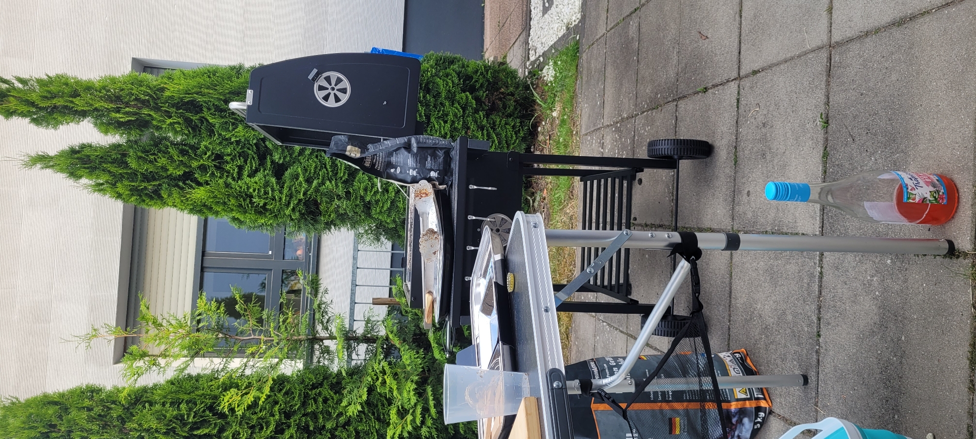 This image depicts an outdoor setting, likely a backyard or patio, in Heusenstamm, Germany. A black charcoal grill is the central focus, with its lid open, revealing a cooking grate. On the grill, a foil pan containing cooked food, possibly burgers or patties, is visible, along with a pair of tongs. To the left of the grill, a portable folding table is set up. On the table, there is a clear plastic cup filled with liquid, a metal tray, and a wooden cutting board. Beneath the table, a bag of charcoal briquettes is placed. In the foreground, a glass bottle with a blue cap, containing a red liquid, stands upright on the paved ground. The background is dominated by a modern building with large windows and several tall, manicured evergreen trees, characteristic of a suburban environment. The ground is paved with square concrete tiles, with patches of grass and gravel visible. No people are present in the scene.