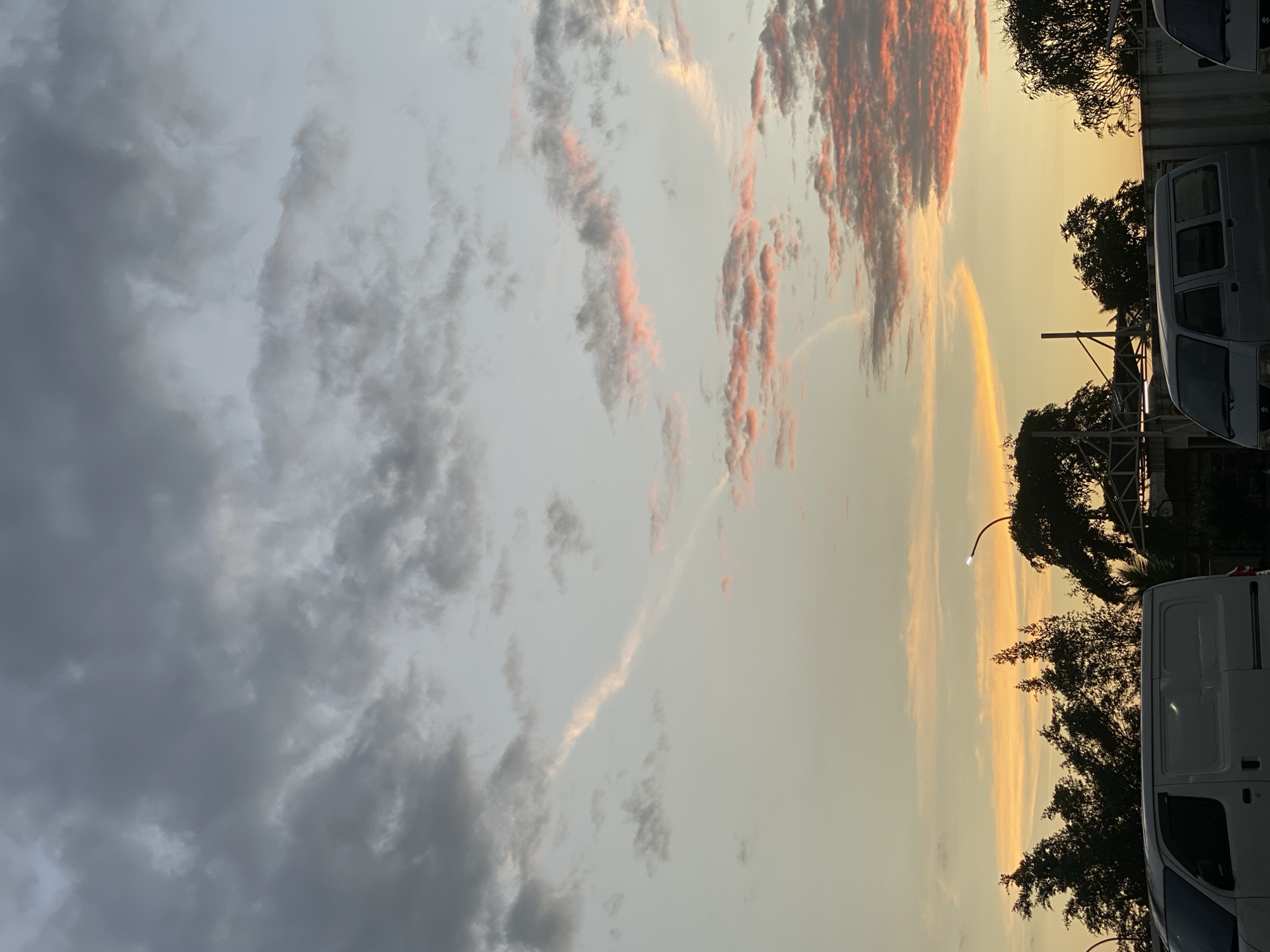 The image captures a sunset in Gambut Barat, Indonesia. The sky is filled with dramatic clouds, with darker gray clouds dominating the upper portion and wispy clouds tinged with pink and orange towards the horizon. Two distinct contrails are visible, one stretching diagonally across the sky and another forming a curved arc near the horizon. Silhouetted against the vibrant sky are the tops of trees, a street lamp, and structures that appear to be part of a residential or industrial area. In the foreground, two white vans are parked, partially obscuring the view of the ground and any potential human activity. The overall impression is one of a peaceful evening scene in a semi-urban or suburban setting.