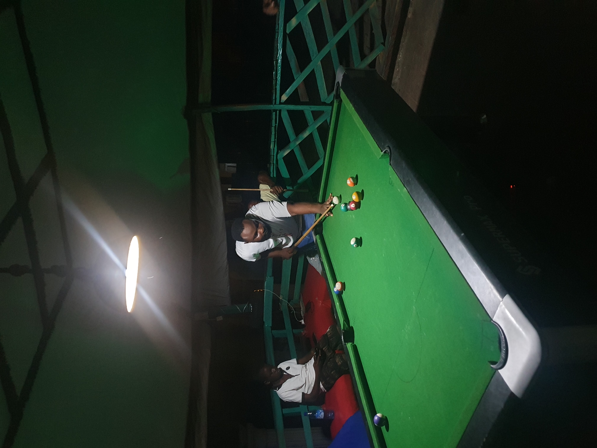 In Lokoja, Nigeria, two individuals are observed at a pool table under a dimly lit, outdoor setting. One man, wearing a white t-shirt and blue jeans, is actively playing pool, cue stick in hand, poised to strike a ball. He is positioned on the right side of the table. To the left, another man, dressed in a white polo shirt and patterned shorts, is seated on a red cushion, looking down at a mobile device. He is partially obscured by the pool table. The pool table itself is green with several colored balls scattered across its surface. In the background, a green lattice fence encloses the area, and a ceiling fan is visible overhead. The overall ambiance suggests a casual recreational space.