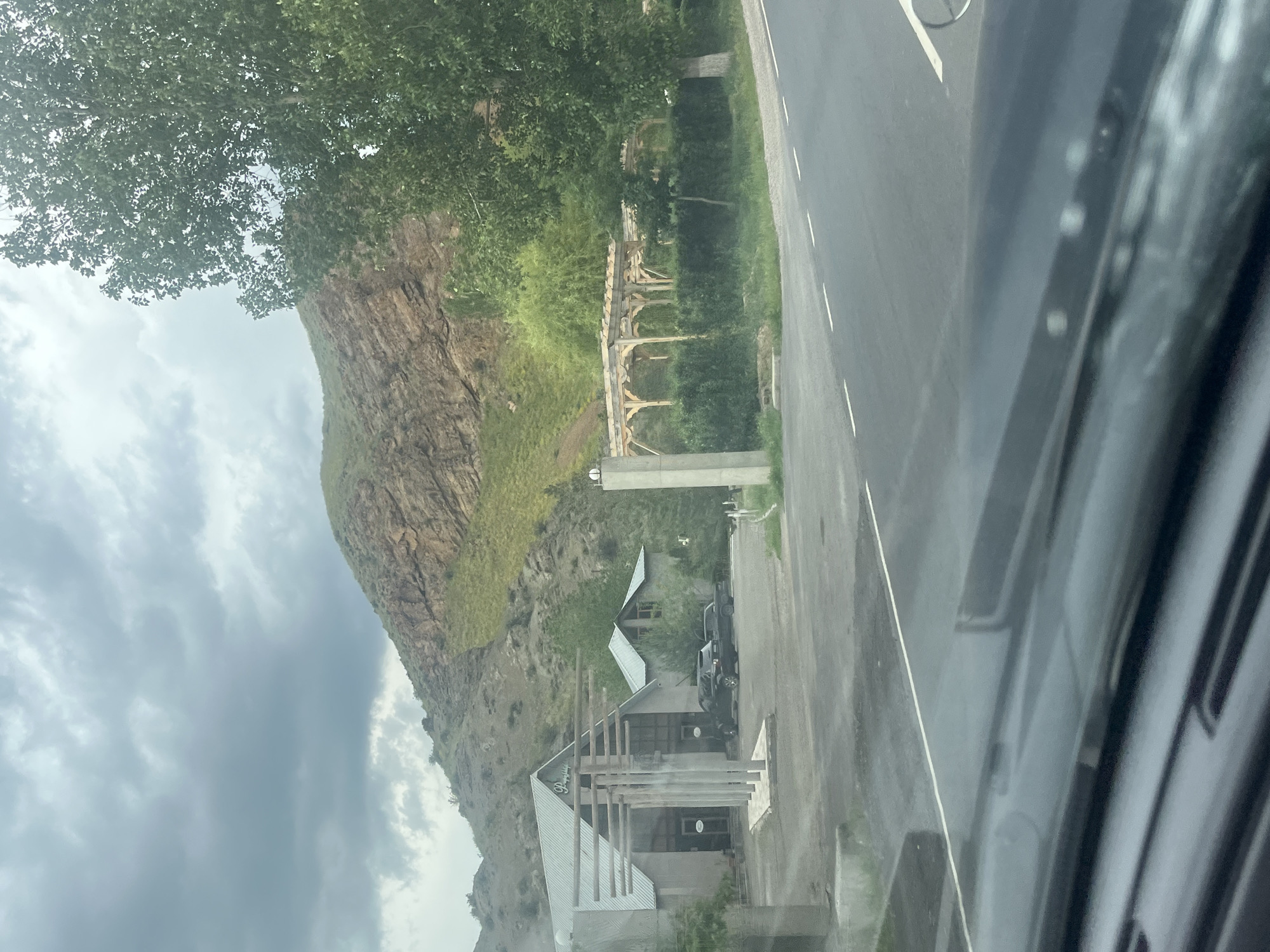 The image captures a rural landscape in Chiva, Armenia, seen from a moving vehicle, with a blurred foreground suggesting a car's interior. The dominant feature is a large, rugged mountain or hill on the left, covered in sparse green vegetation that gives way to a rocky, textured face. Dense deciduous trees crown the upper slopes, casting shadows.
A paved road with white double solid lines and dashed markings runs horizontally across the right side of the frame. Alongside the road, a line of well-maintained hedges separates it from a series of modern buildings and structures. One prominent building, likely a business, features a distinctive angular metal roof and a façade adorned with vertical wooden beams forming an entryway. Parked nearby are at least two dark-colored pickup trucks or SUVs. Further back, another similar building is visible.
A notable architectural element is a wooden pergola-like structure with arched supports, situated amidst lush greenery between the road and the buildings. The sky above is overcast with scattered grey clouds, indicating a cloudy daytime. No people are visible in the frame.
On the main building, a partially visible sign displays text. The letters are light-colored against a dark background, clearly showing "Poyraz" in a stylized font, followed by what appears to be "Shop" or "Hop," suggesting a business establishment like a store or restaurant. The scene conveys a blend of natural ruggedness and modern rural development.