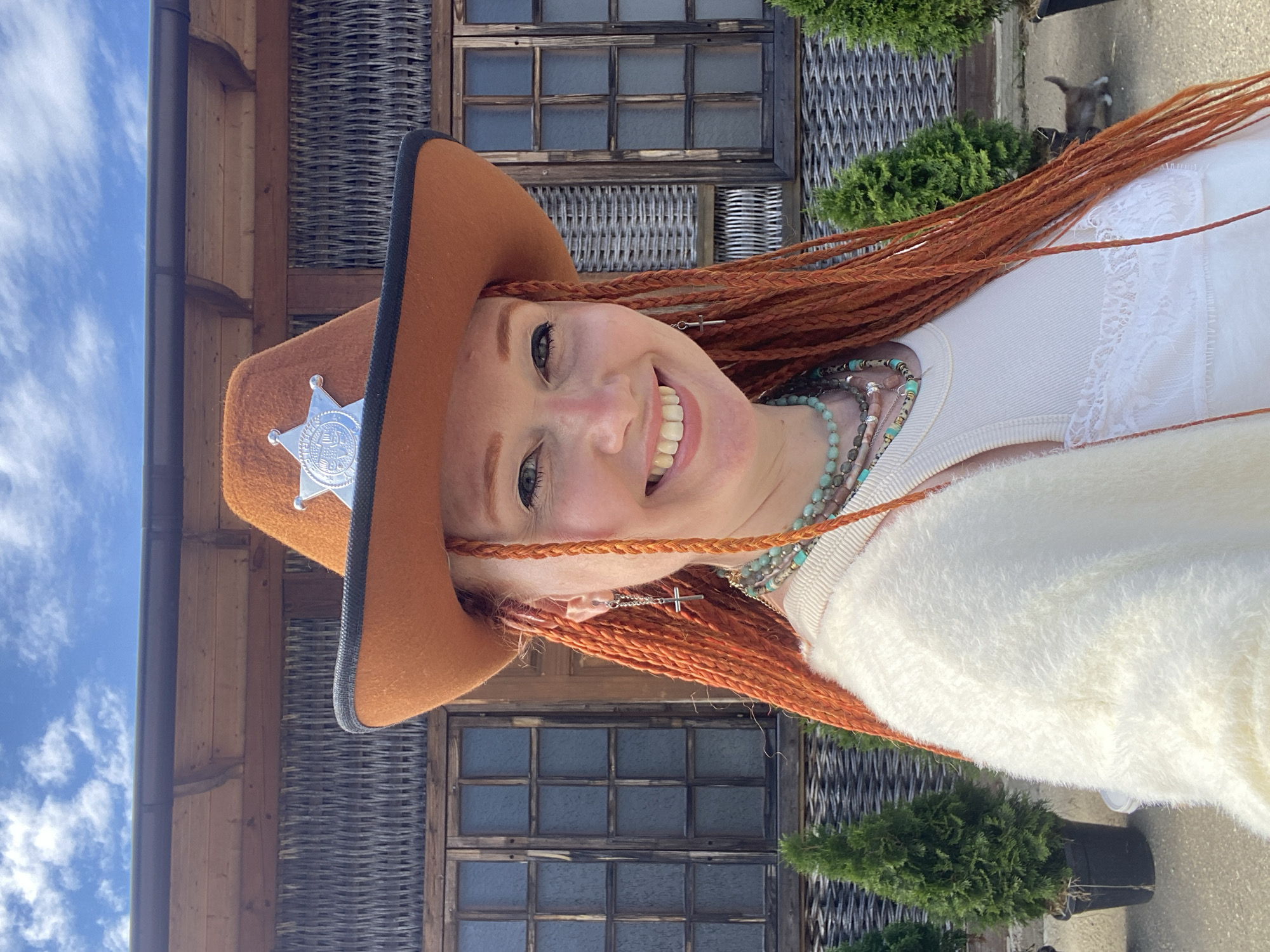 A woman with long, bright orange braided hair and fair skin wears a brown cowboy hat with a silver sheriff's badge. She is smiling and wearing a white tank top with lace trim and a fuzzy white cardigan. She has on multiple necklaces with beads and charms, including a cross pendant. The background features a wooden building with woven detailing and windows, as well as several small evergreen trees in black pots. The sky above is blue with white clouds. A small kitten is visible walking on the ground in the background. The location is identified as Sochi, Russia.