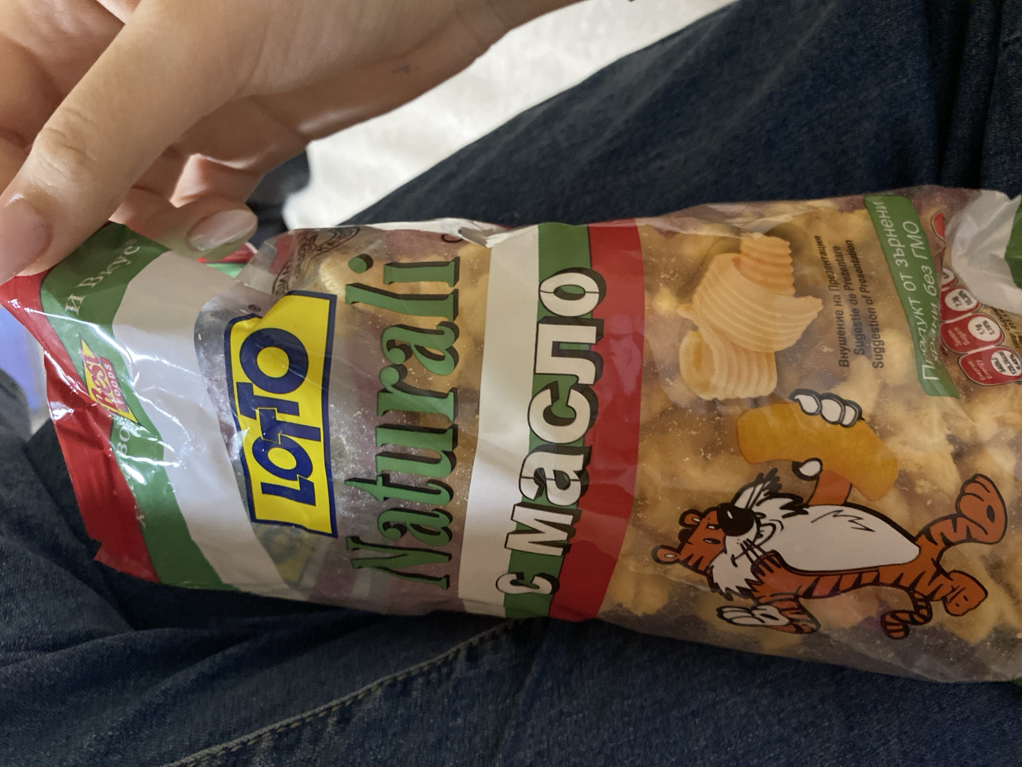A close-up shot shows a person's hand holding a transparent bag of "Lotto Naturali" brand snacks, possibly crisps or puffed edibles, against a background of denim jeans. The person's hand has light-colored manicured nails. The bag contains light-colored, irregularly shaped snacks, some resembling butter curls.
The bag's label features a red, white, and green striped design. Prominently displayed are the words "LOTTO Naturali" and "с масло" (with butter), indicating the flavor. A cartoon tiger character, wearing sunglasses, is depicted on the bag, holding a piece of the snack. Other visible text includes "Внушение на Презентация / Sugestie de Prezentare / Suggestion of Presentation" in Bulgarian, Romanian, and English, along with "Продукт от зърнени храни без ГМО" (Product from cereal grains without GMO). Partial nutritional information is visible, showing "Енергия / Energy" with values "540kJ / 129,3kcal" and "Захари / Sugars" at "1,19%". The words "Rest FOODS" and "Вкус" (Taste/Flavor) are also partially visible on the top left. The setting appears to be casual and indoors, with the person likely seated.