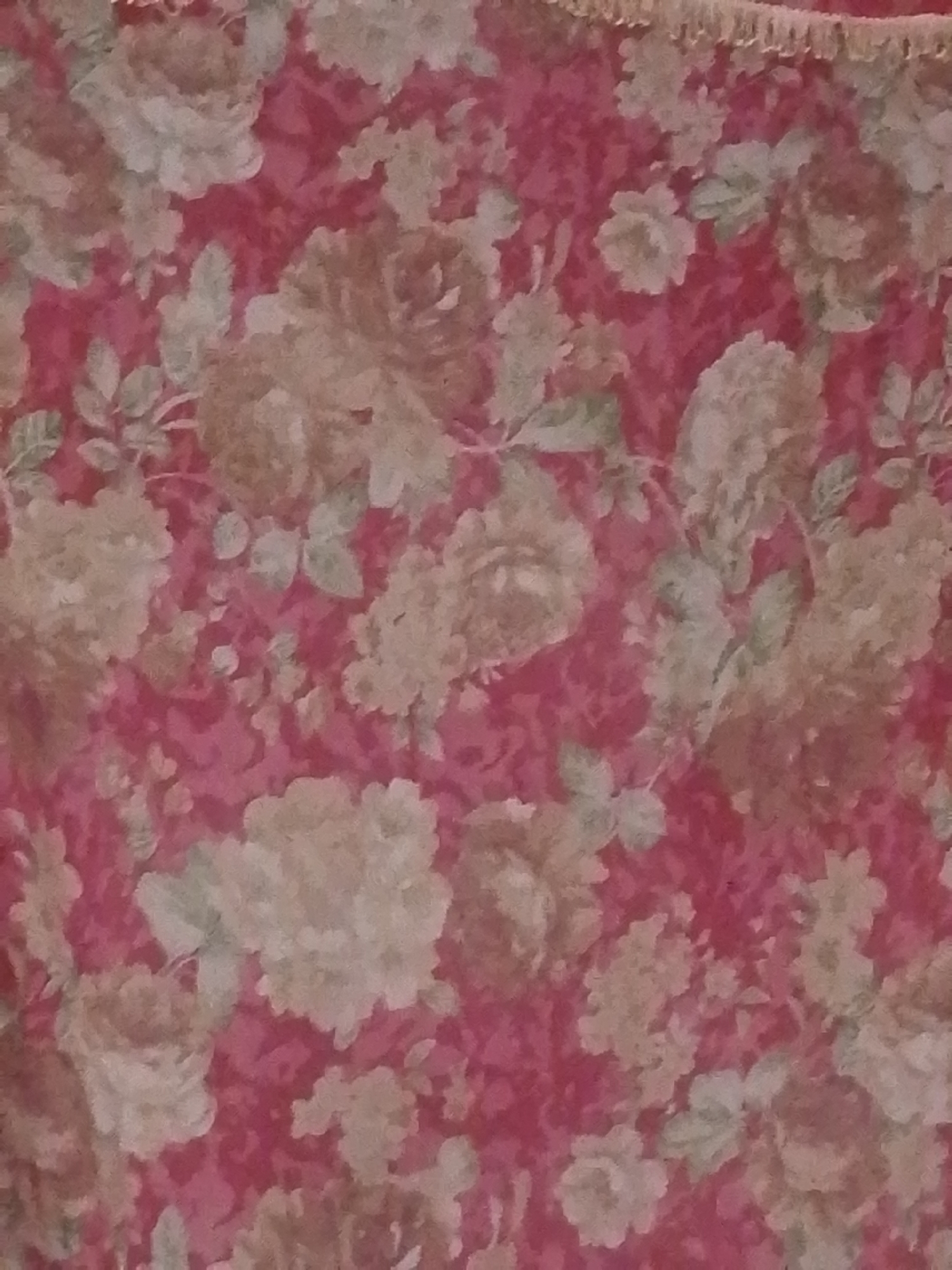 The image depicts a close-up view of a floral patterned fabric. The pattern consists of large, stylized roses in shades of cream and light brown, set against a vibrant pink background. Green leaves are interspersed among the flowers, adding a touch of natural contrast. The fabric appears to be textured, with a slightly raised quality to the floral elements. The top edge of the image shows a hint of fringe, suggesting it might be part of a curtain, tablecloth, or upholstery. There is no discernible location in the city of Abuja, Nigeria, as the image is focused on the fabric's pattern and texture, not on any specific environment or setting.