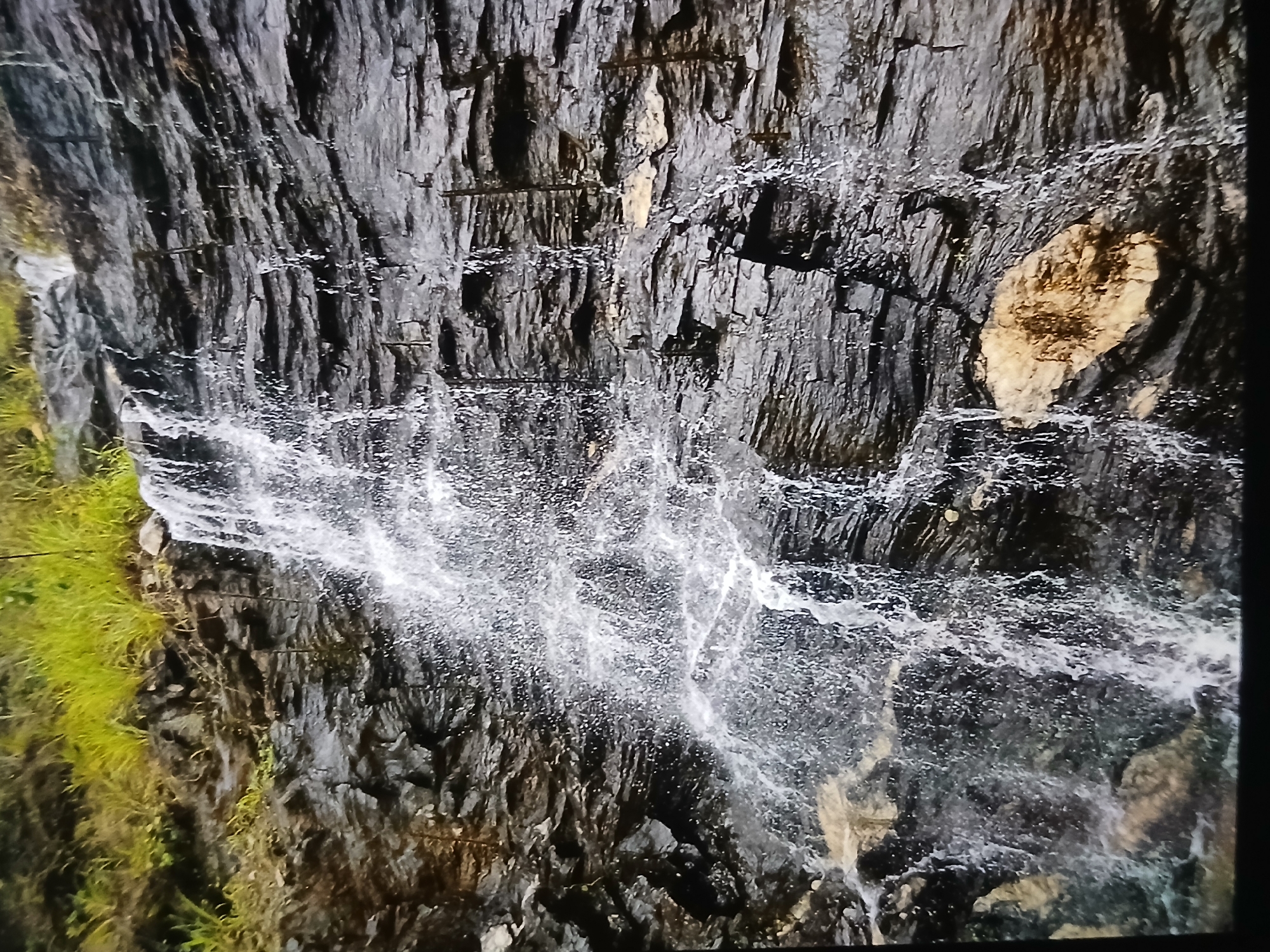 A close-up shot captures a waterfall cascading down a dark, rugged rock face. The water is a blur of white against the dark grey and black of the rocks. Patches of green vegetation cling to the rock on the left side of the frame. There is an isolated patch of light-brown rock on the top right side of the waterfall. The location is not clearly visible, and therefore it cannot be determined whether the image is from Bengaluru, India.