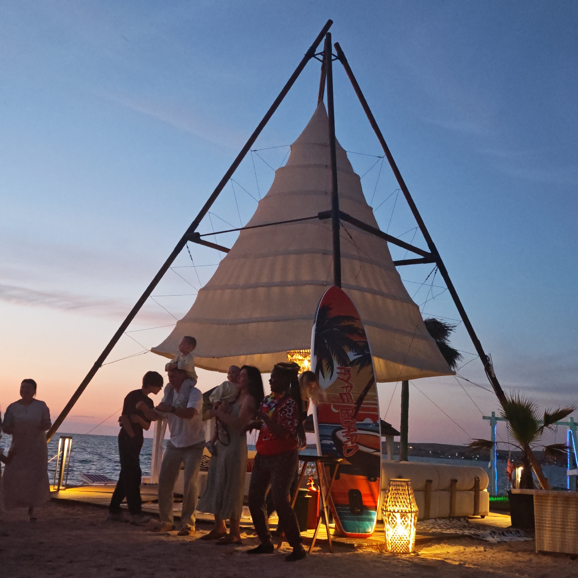 At dusk on a sandy beach, a group of people are gathered near a tent-like structure. A child is being carried on someone's shoulders. A surfboard leans against the structure. In the background, the sea is visible, with a faint orange and pink gradient in the sky. There are also decorative lamps and furniture present. The scene suggests a relaxed outdoor gathering or event.