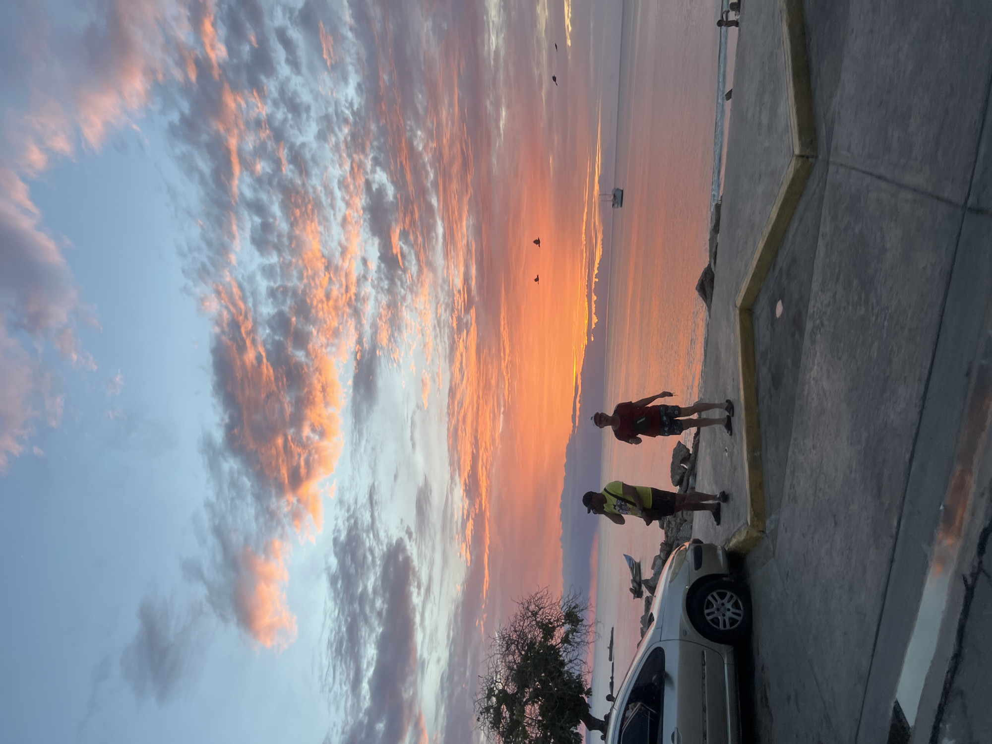 At sunset in Juan Griego, Venezuela, two men stand on a concrete embankment overlooking the sea. The sky is ablaze with vibrant hues of orange, pink, and blue, as clouds are illuminated by the setting sun. A silver car is parked on the left side of the frame. In the distance, a boat is visible on the water, and a larger ship sails further out. Several birds are in flight in the sky. The overall scene captures the natural beauty of a coastal sunset in Venezuela.